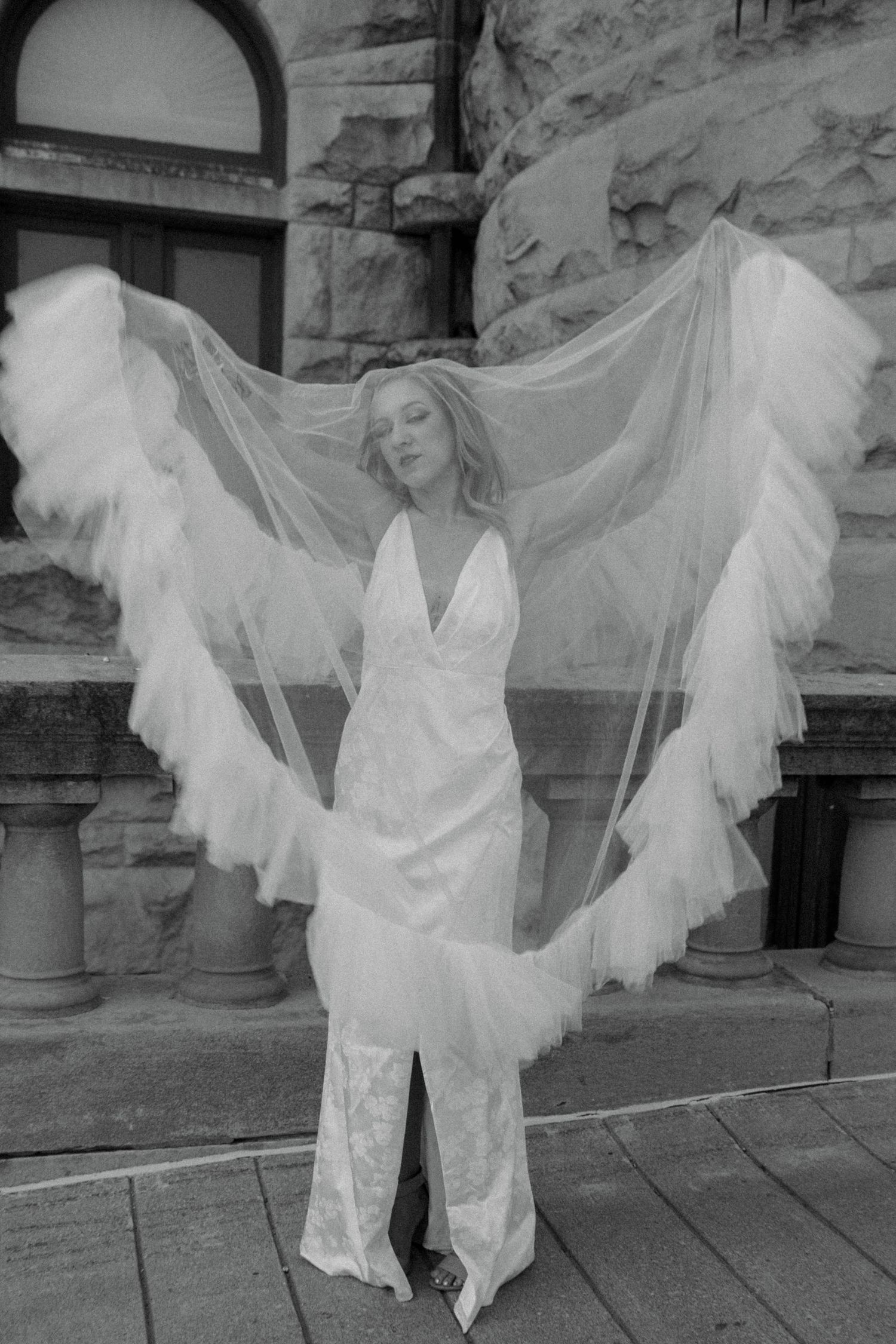 Bride in fitted lace gown with large ruffled veil spread like wings in black and white photo at stone building.