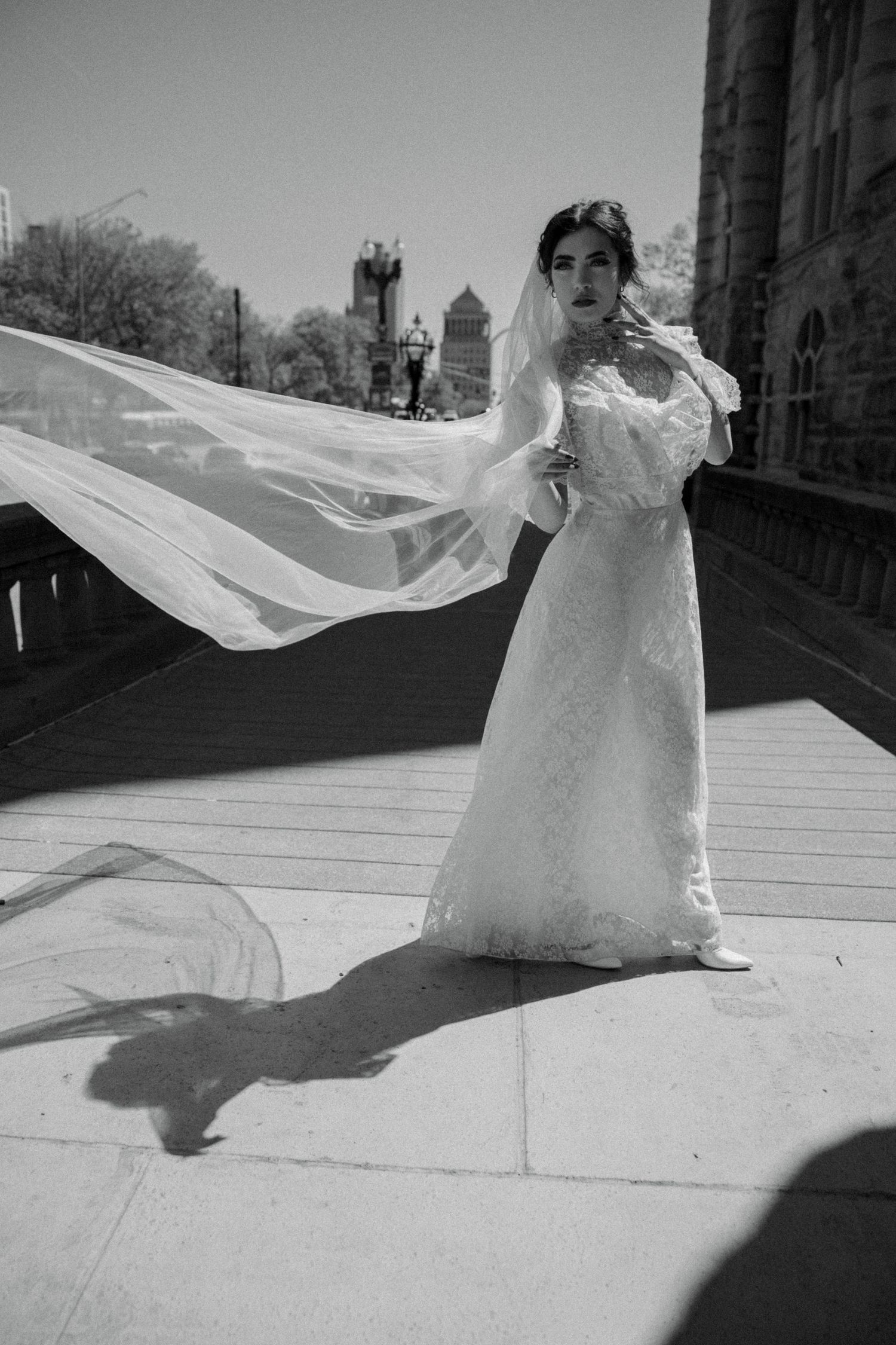 Bride in lace wedding gown with flowing veil caught by wind on urban bridge, black and white photo.