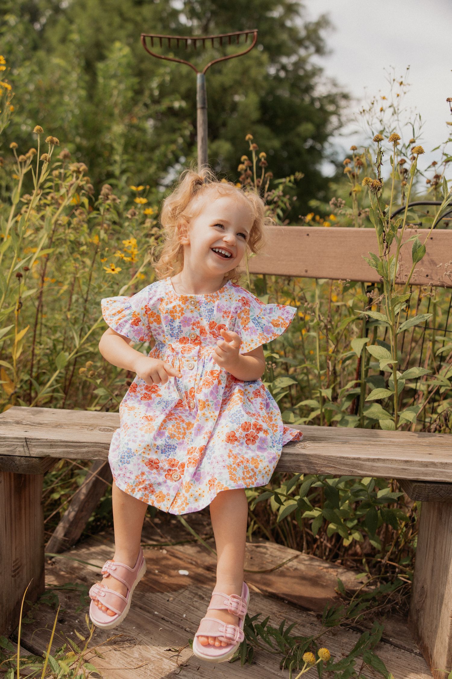 Child in floral dress laughs while sitting on rustic wooden bench in garden setting.