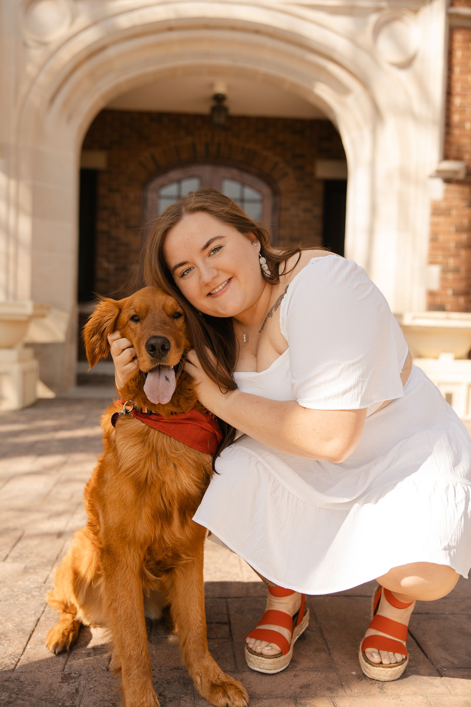 A happy Golden Retriever with a red collar poses with someone in white beside a brick building.