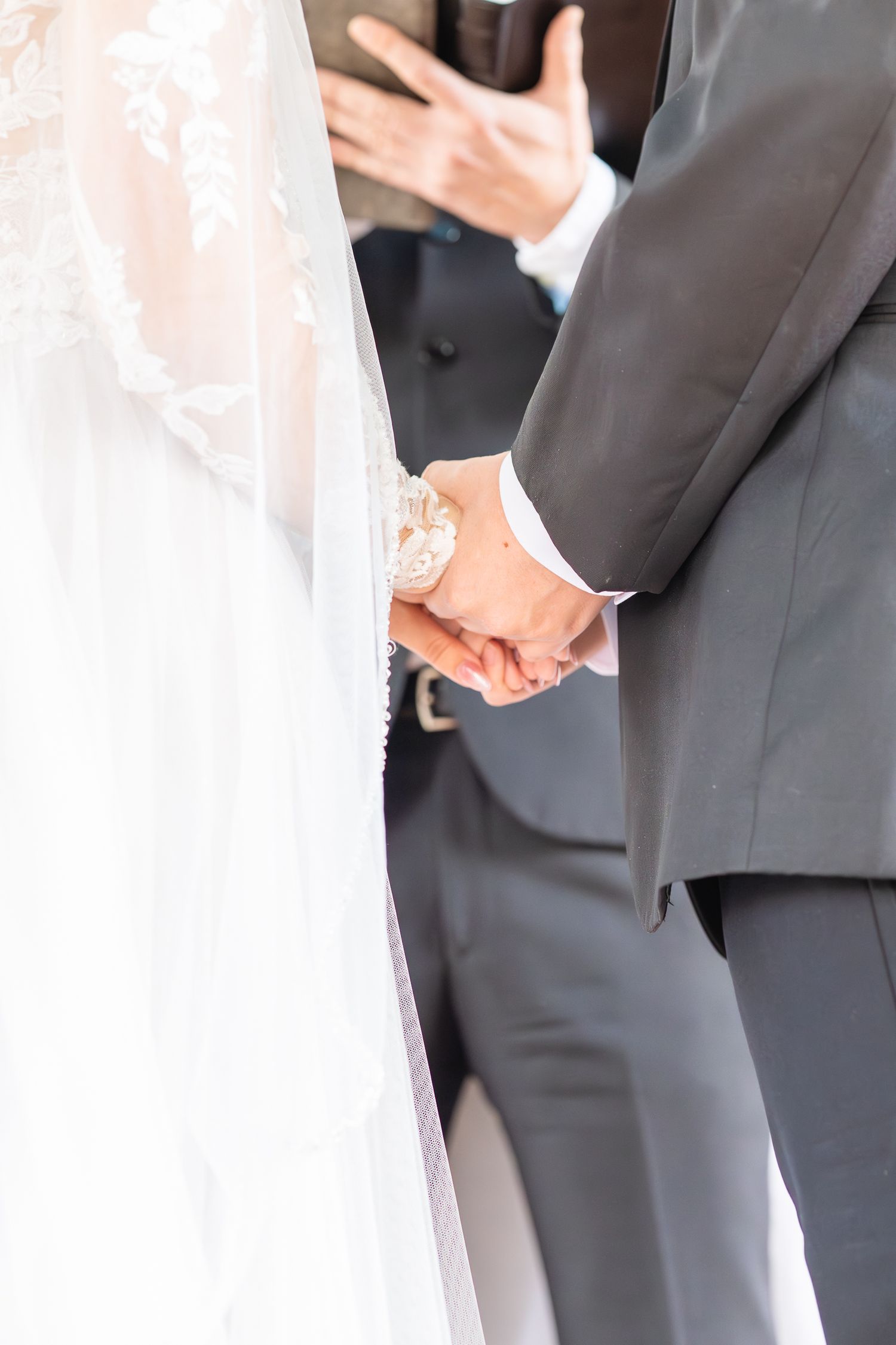 Close-up of couple holding hands during wedding ceremony, with white veil detail.