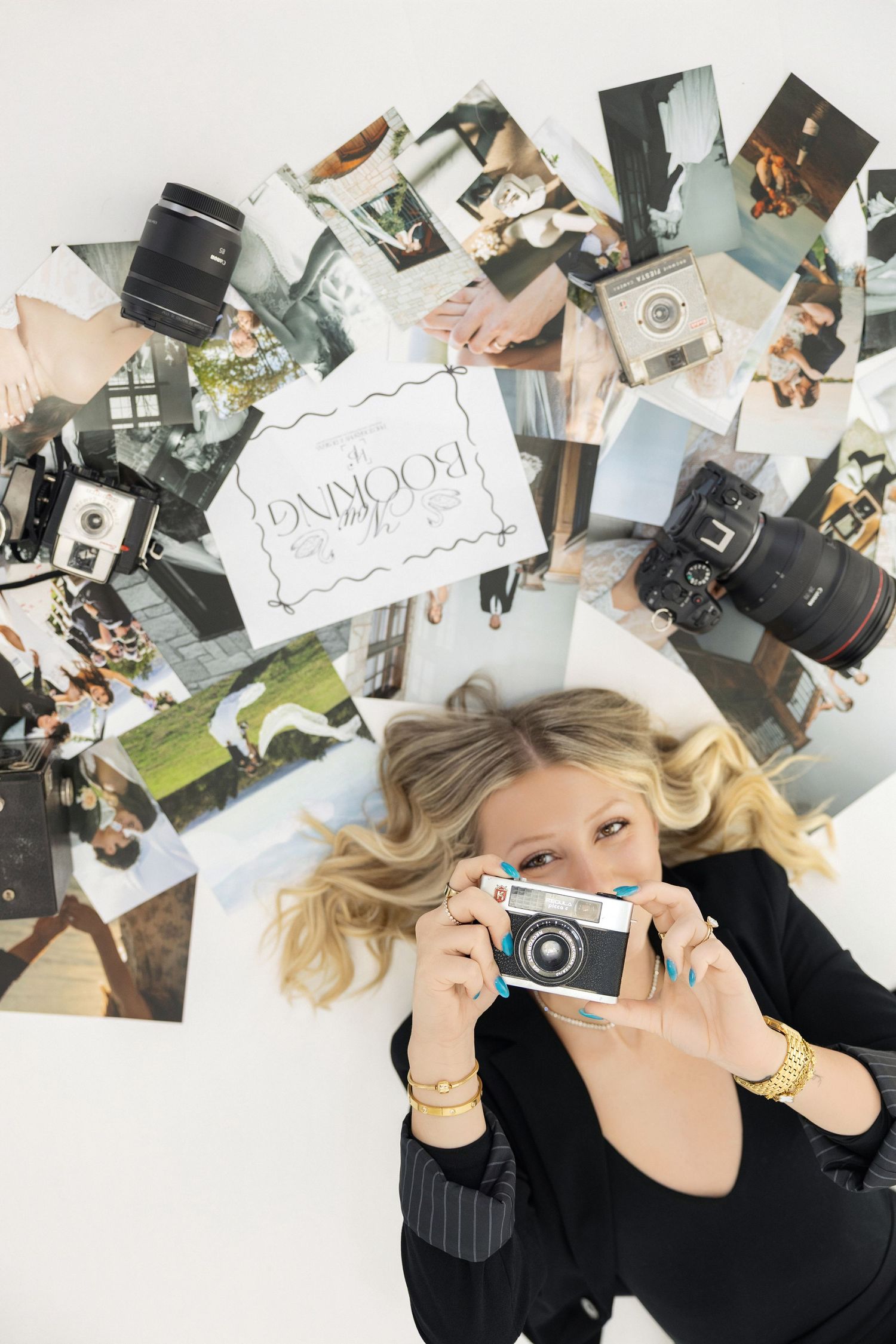 Blonde woman lying on floor holding vintage camera surrounded by photography prints, lenses, and a booking sign from above.