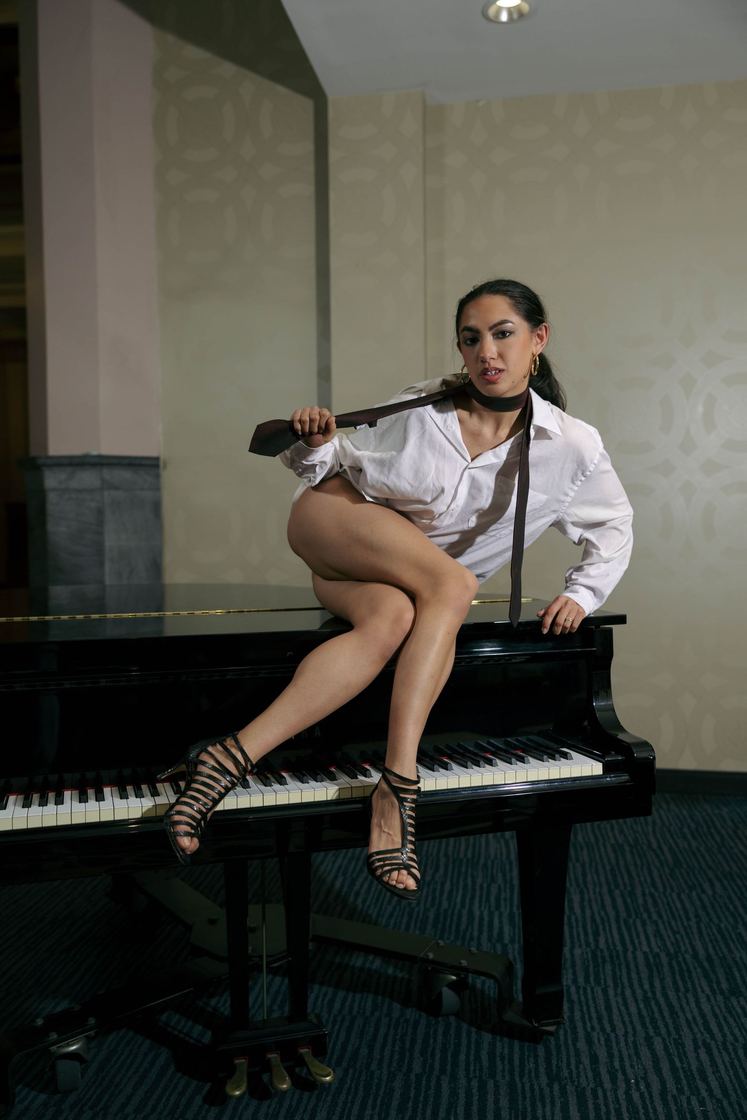 Woman in white shirt and heels sitting on a grand piano, holding a tie, in an elegant room.