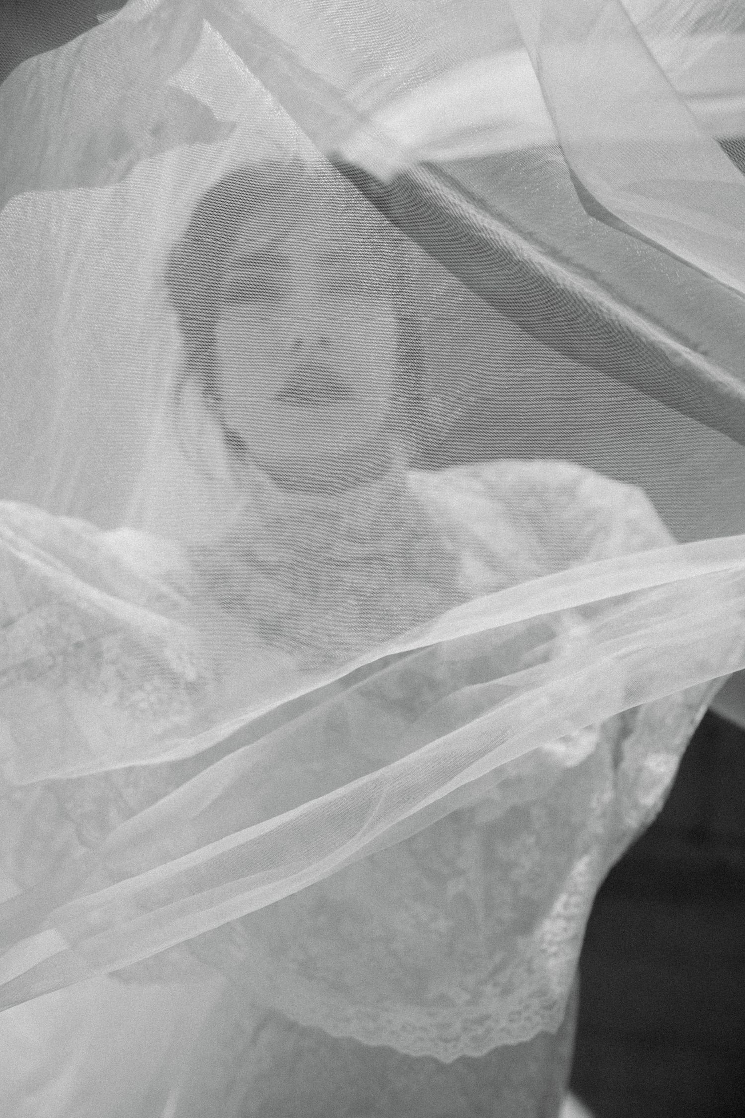 Black and white photo of a bride in lace gown, her face softly visible through flowing wedding veil.