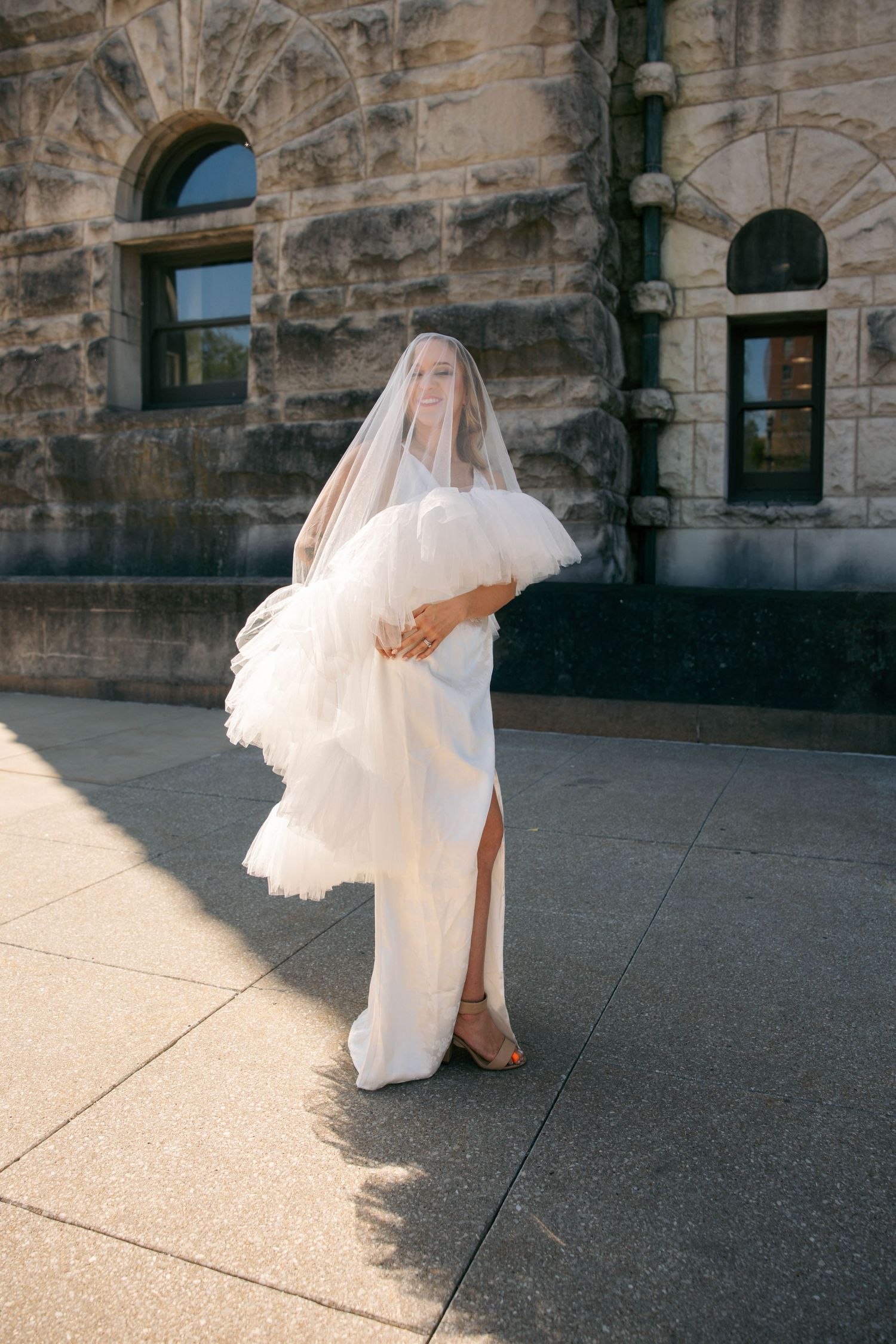 Bride in sleek white gown with dramatic tulle sleeves and veil smiles in warm sunlight by stone building.