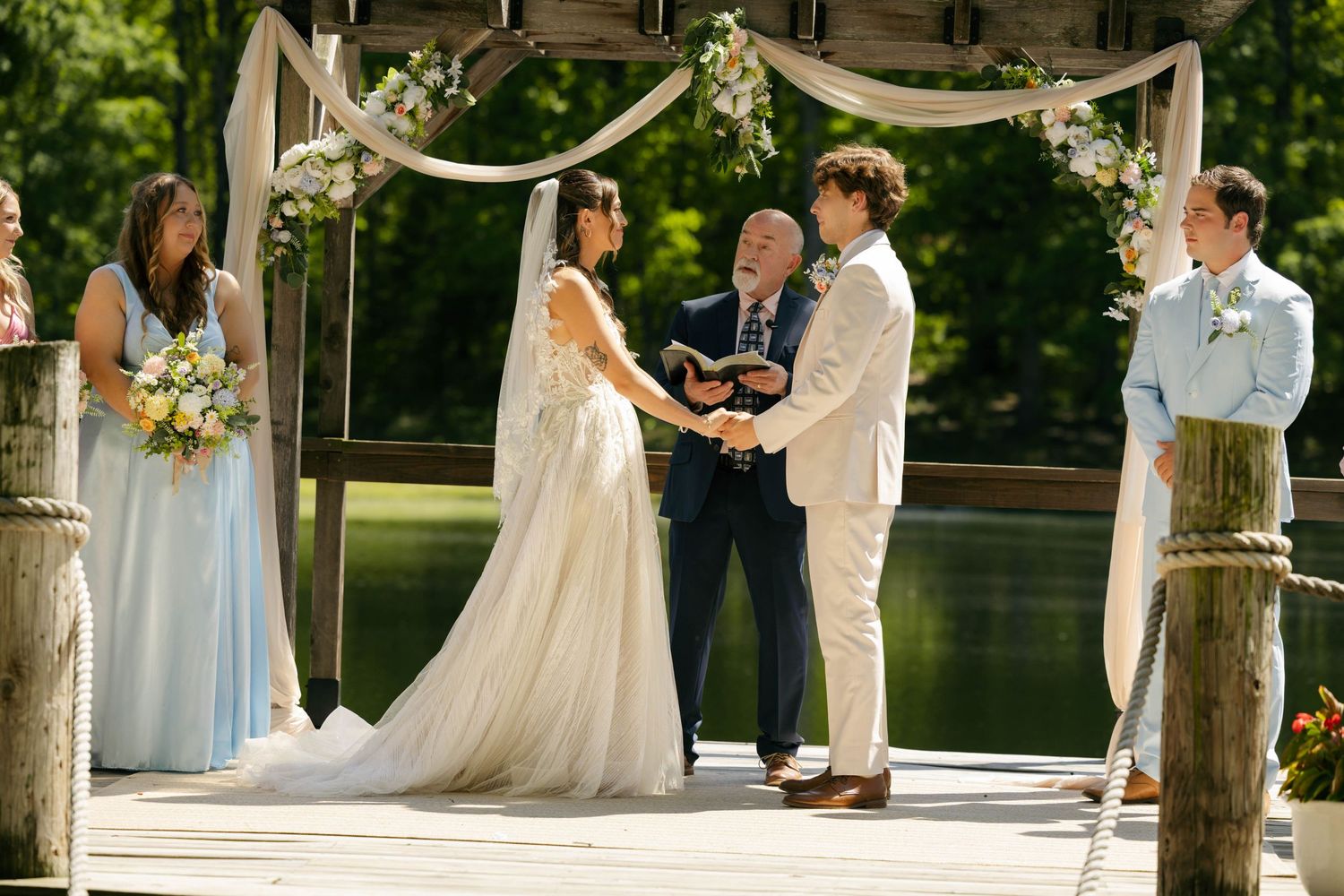 Bride and groom hold hands at outdoor waterfront wedding ceremony under floral arch on wooden dock.