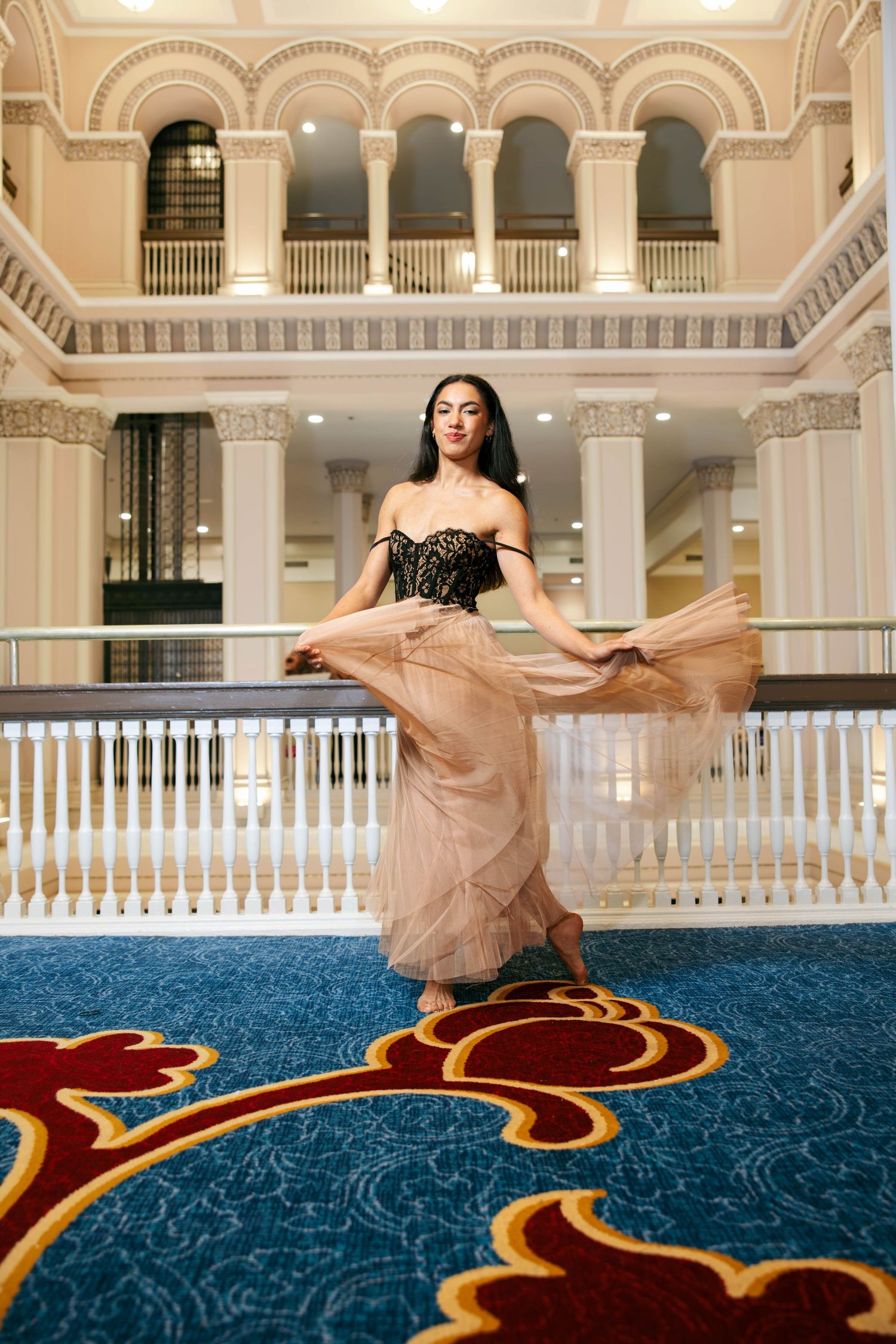 Elegant woman in black corset and flowing beige tulle skirt posing in a grand ballroom with ornate architecture.