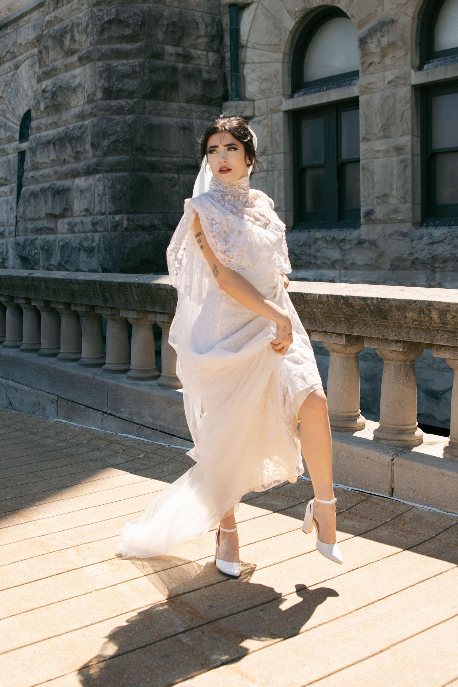 Bride in white lace wedding dress and veil walking on stone terrace of historic building in sunlight.