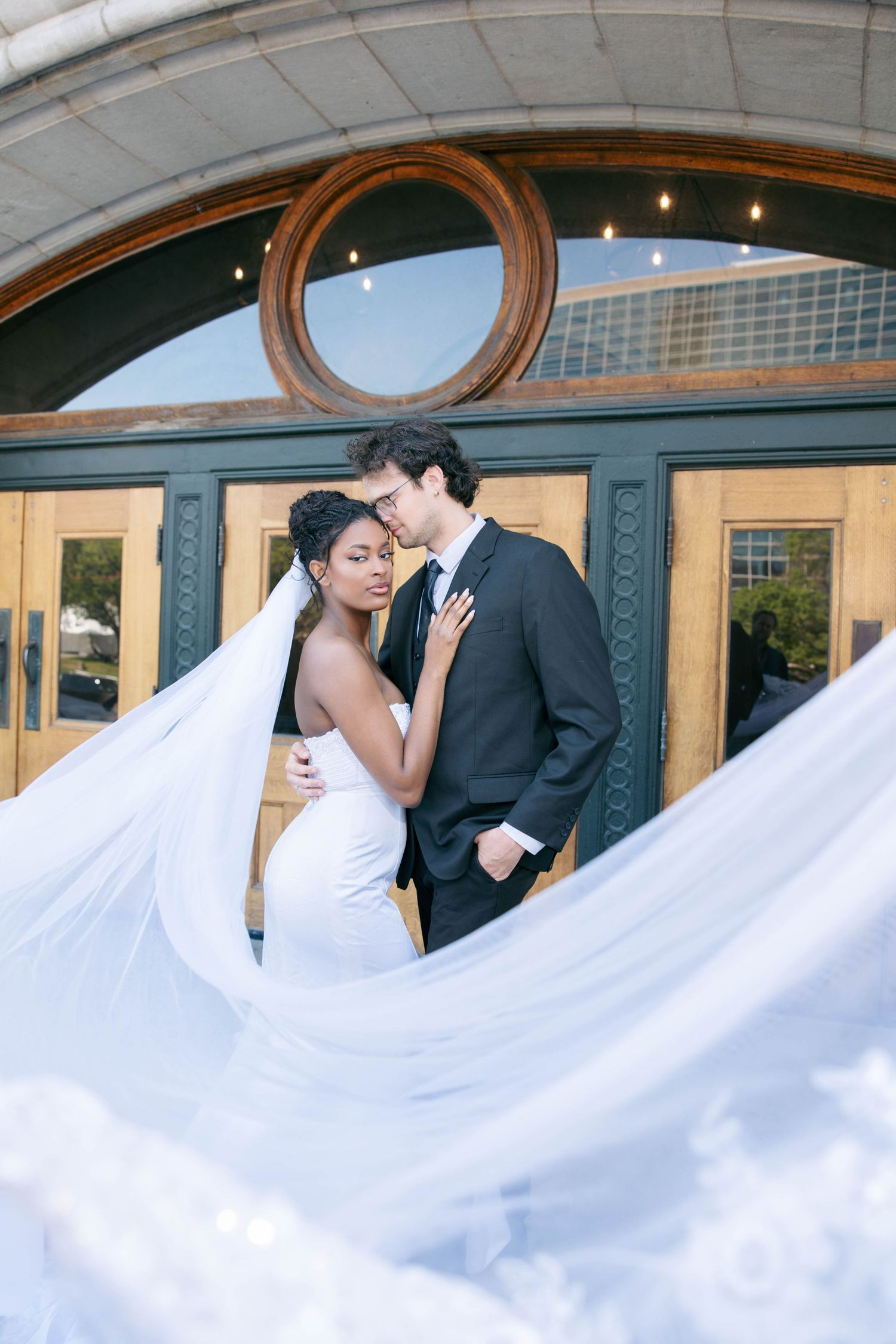 Interracial couple embracing romantically with flowing veil billowing around them outside elegant venue doors.