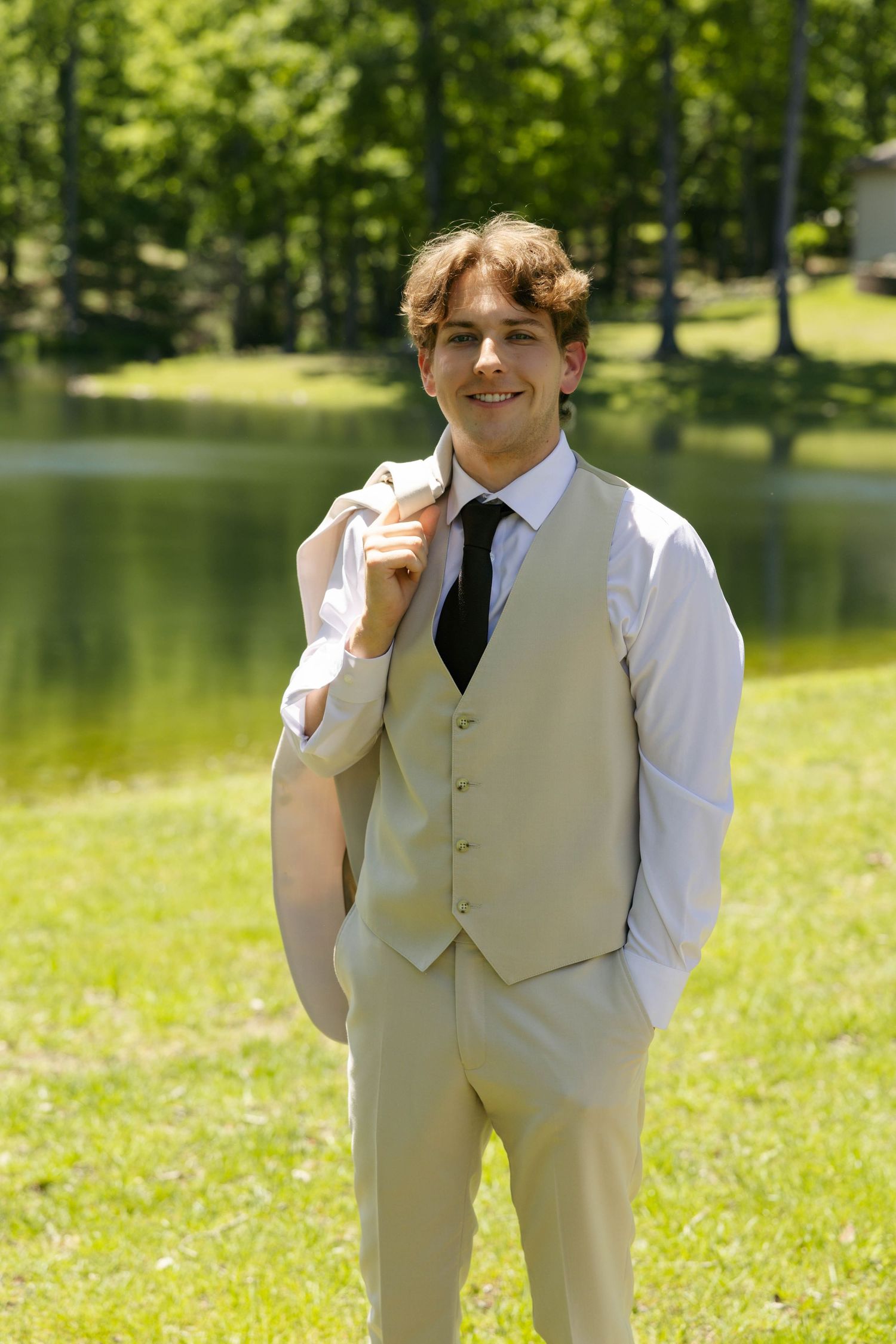 Young man in beige vest and black tie holding jacket over shoulder, smiling outdoors near a pond.