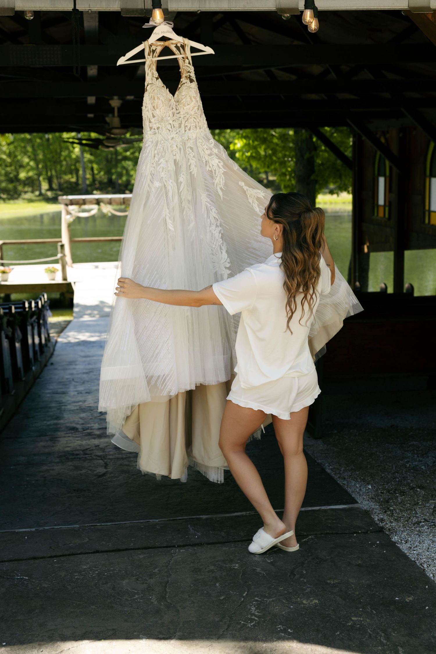 Bride in white robe admiring her lace wedding dress hanging in a rustic outdoor pavilion surrounded by greenery.
