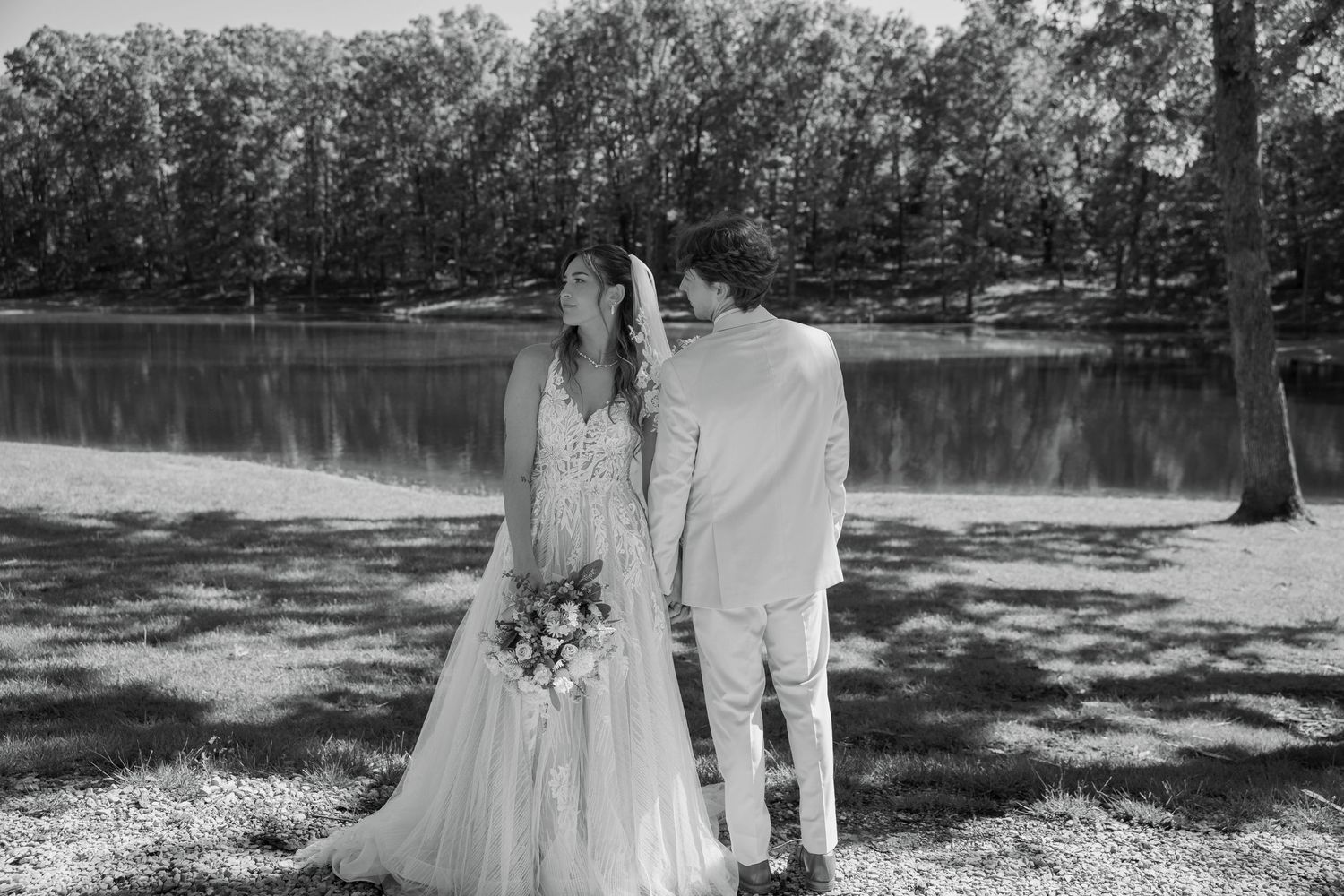 Black and white photo of a bride and groom holding hands by a lake, surrounded by trees on their wedding day.