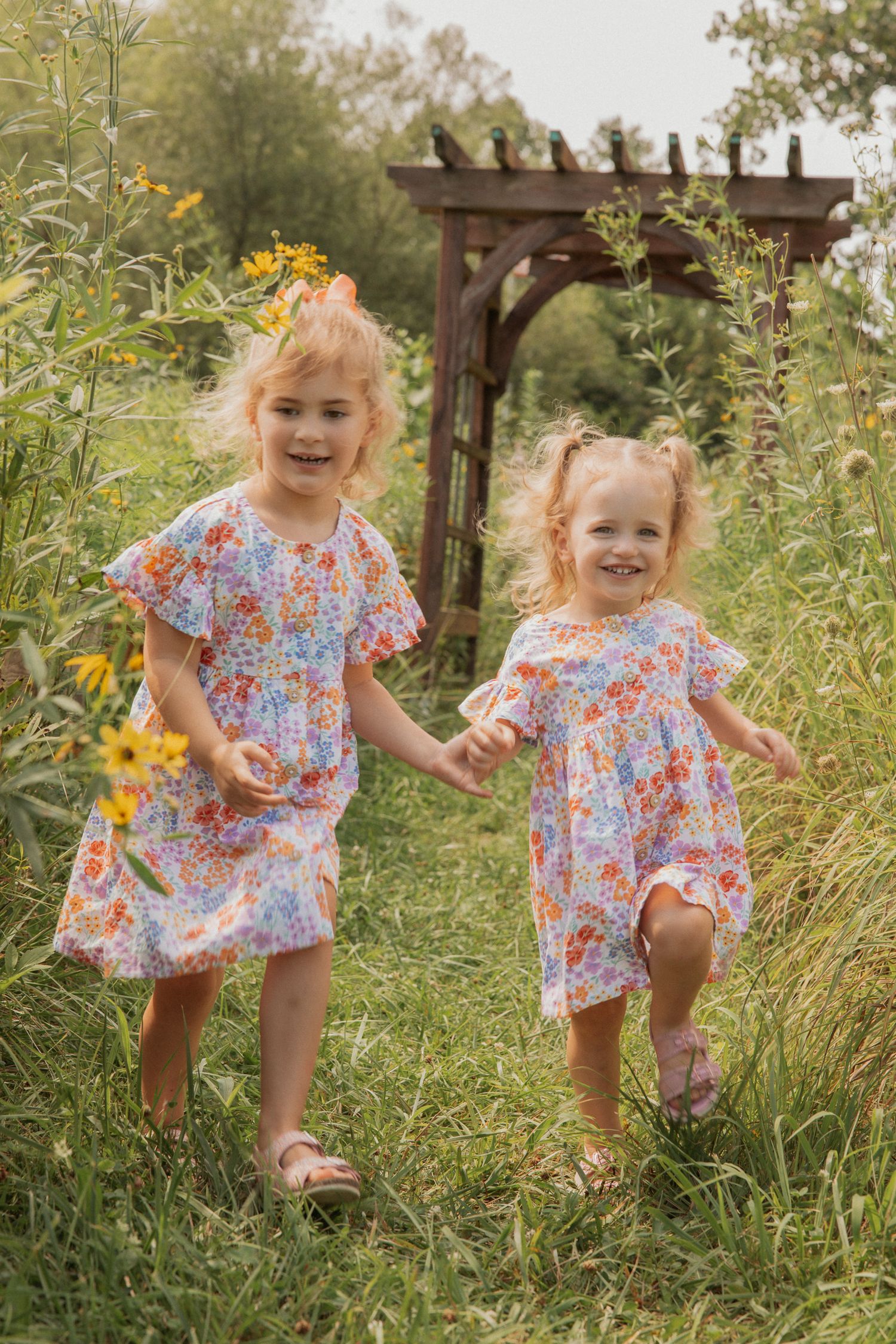 Two children in matching floral dresses laughing and playing by wooden pergola.