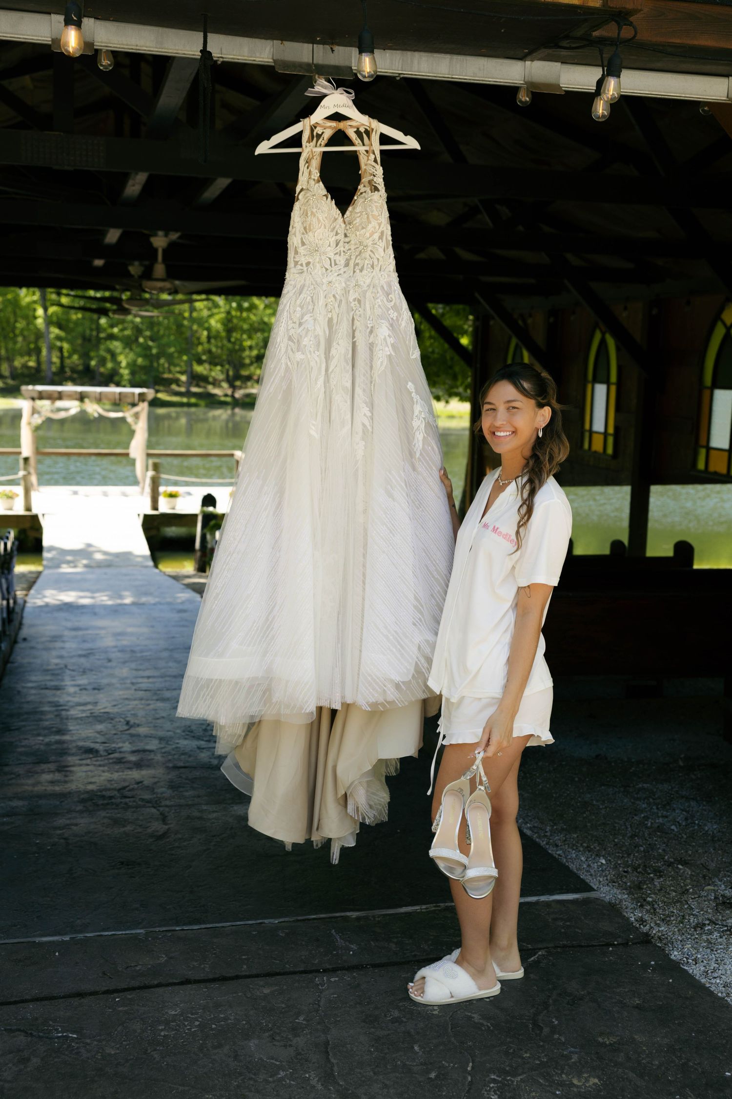 Smiling bride in white robe holding wedding shoes beside her elegant lace halter wedding gown hanging in a rustic venue.