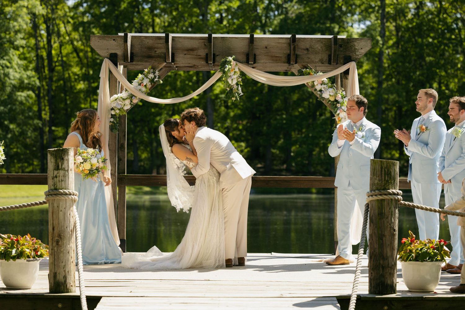 Bride and groom share first kiss at outdoor lakeside wedding ceremony under draped floral arch on dock.