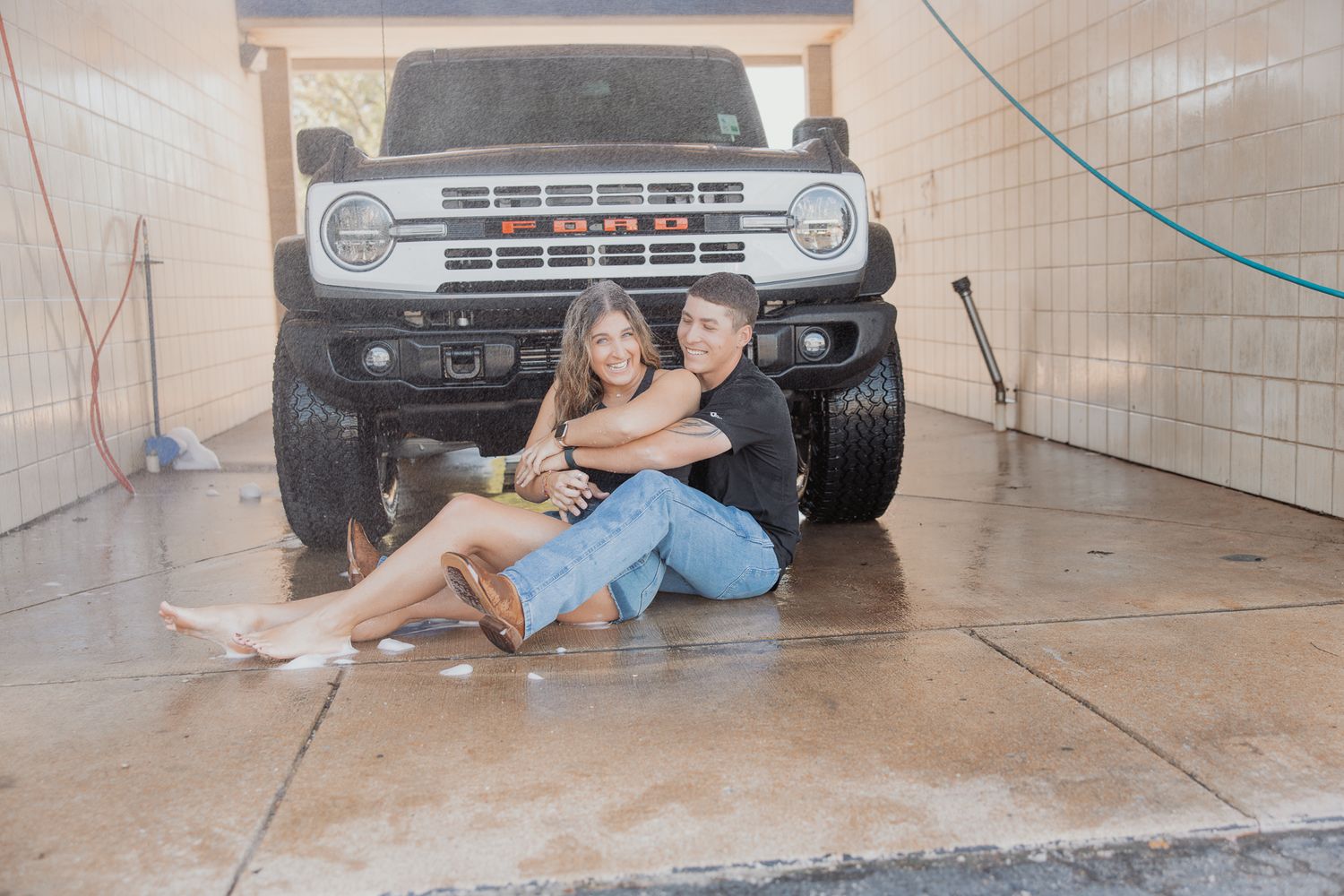Couple sits together on garage floor with their dog in front of a lifted Ford truck.