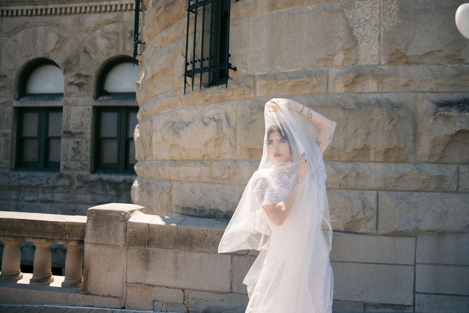 Bride in white wedding dress and veil standing outdoors near stone building balustrade on sunny day.