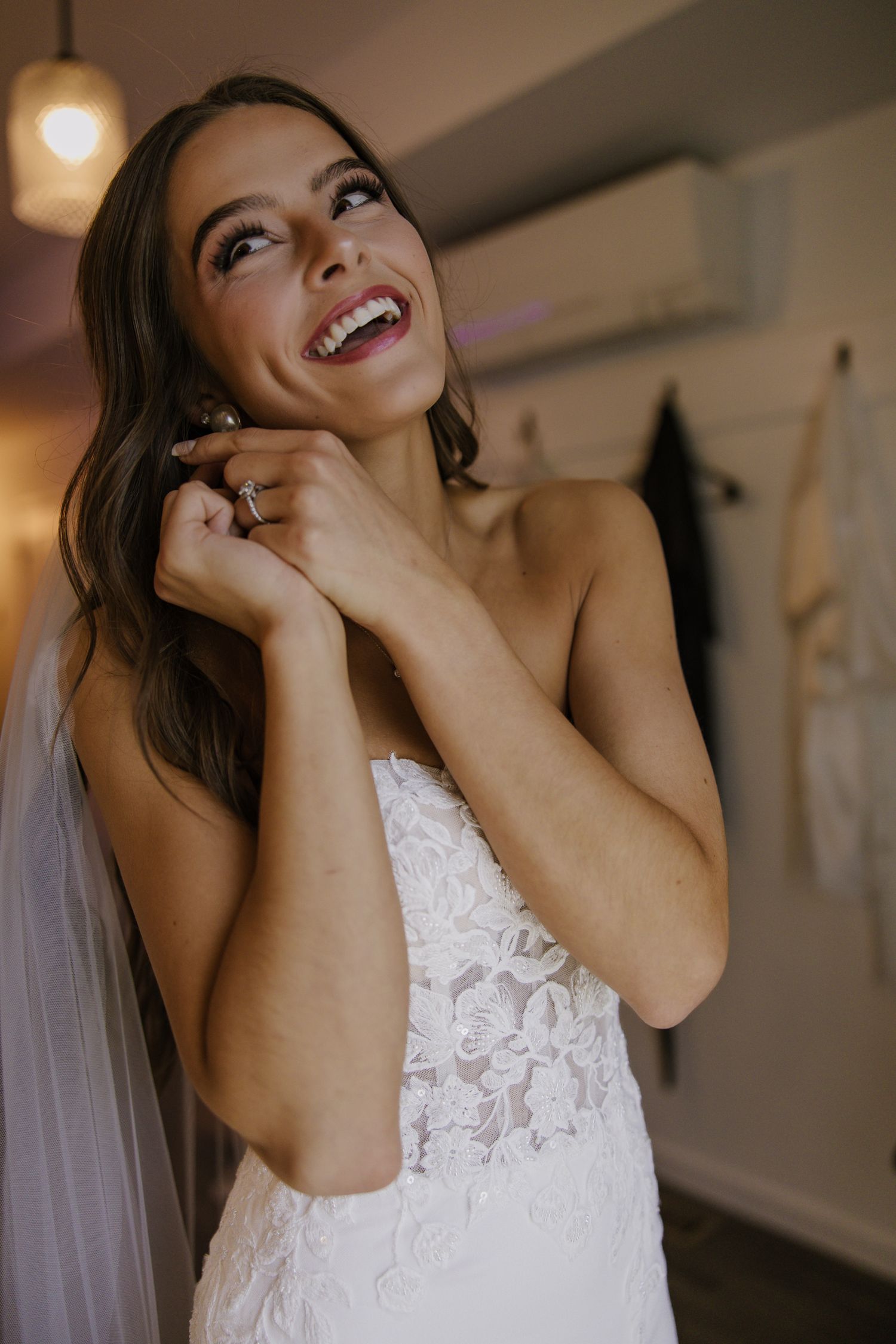 Smiling bride in lace wedding dress adjusting earring while wearing veil in bridal suite.
