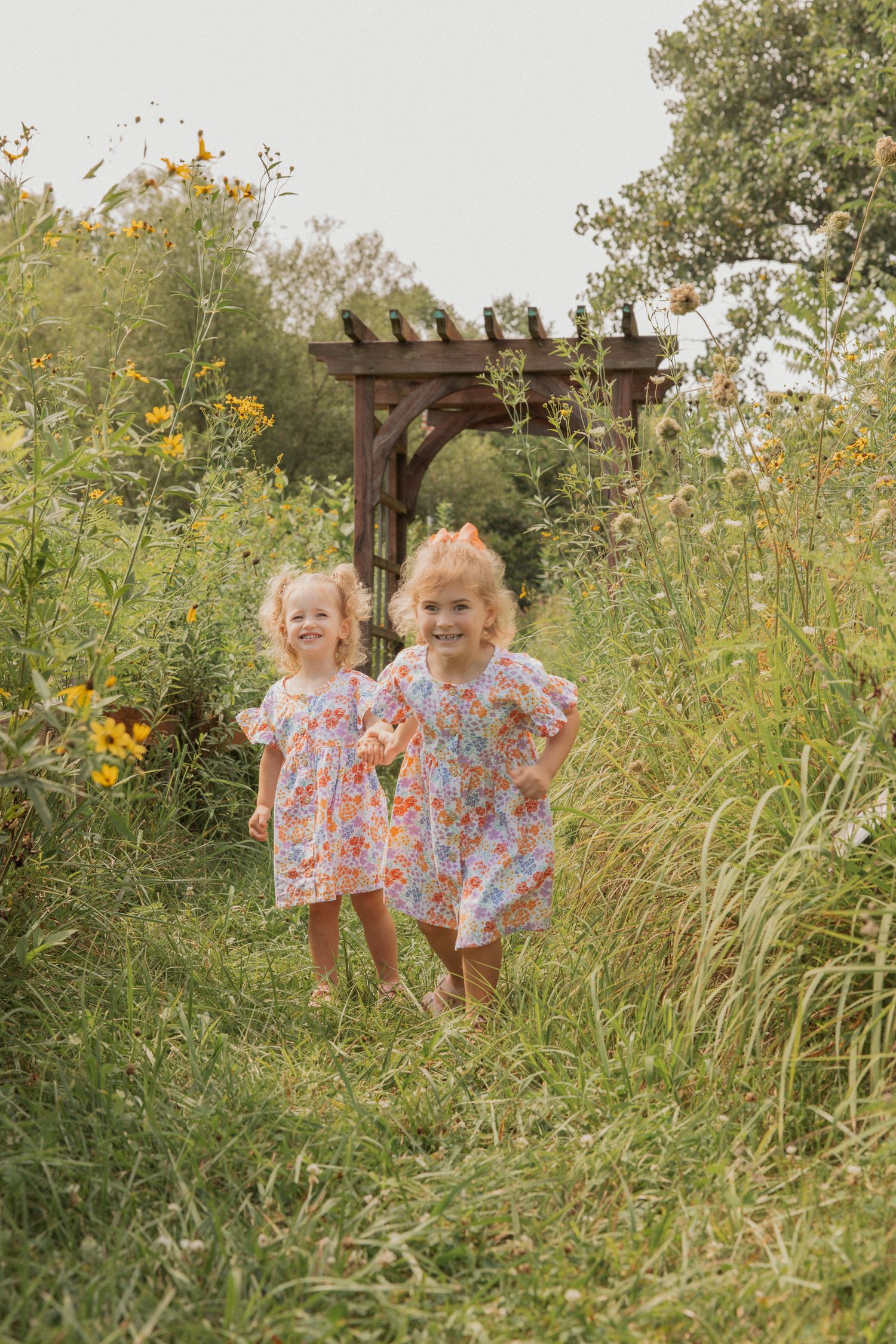Two children in matching floral dresses standing by wooden pergola in wildflower field.