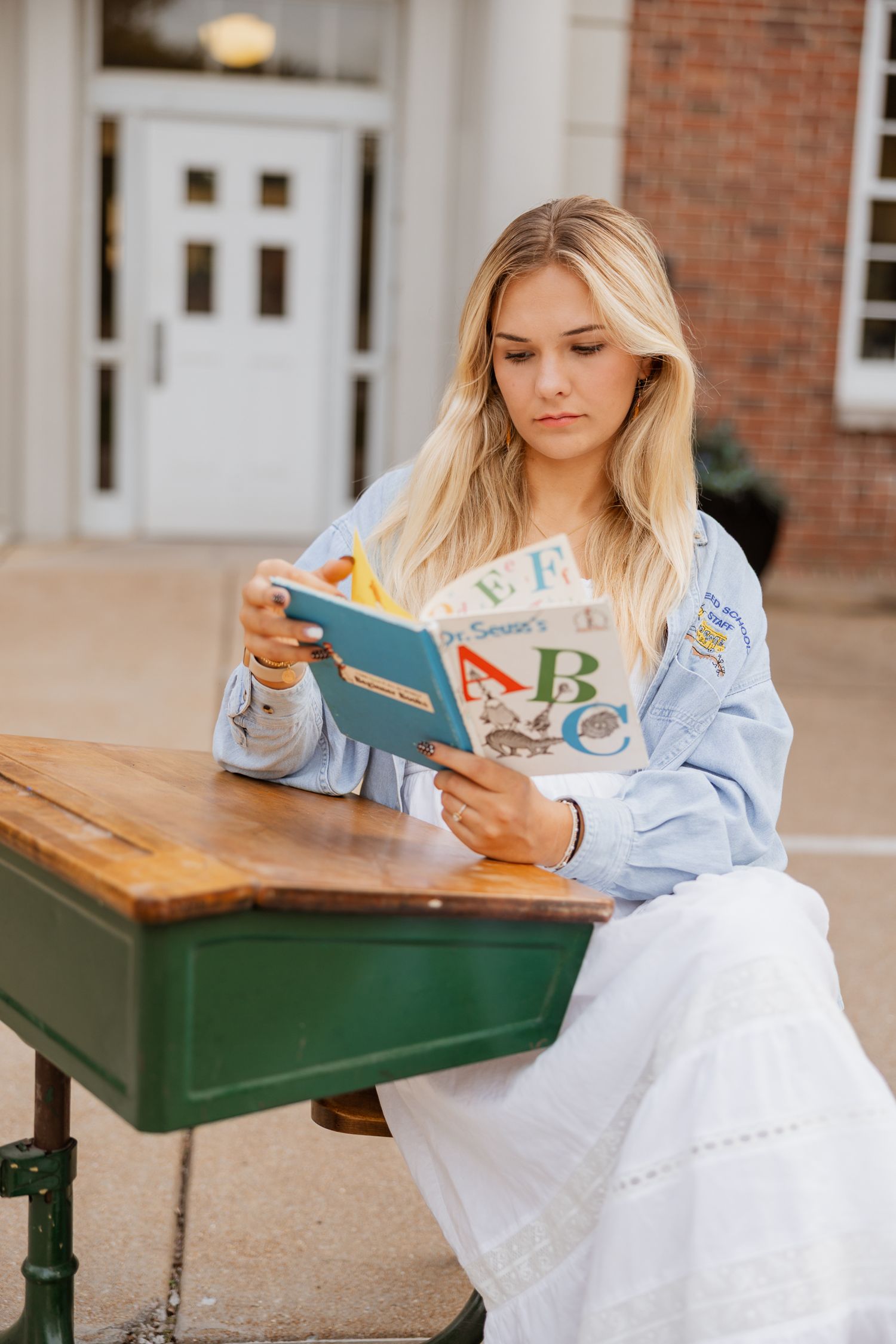 A person in a blue shirt and white dress reads a children's book while sitting at an outdoor table.