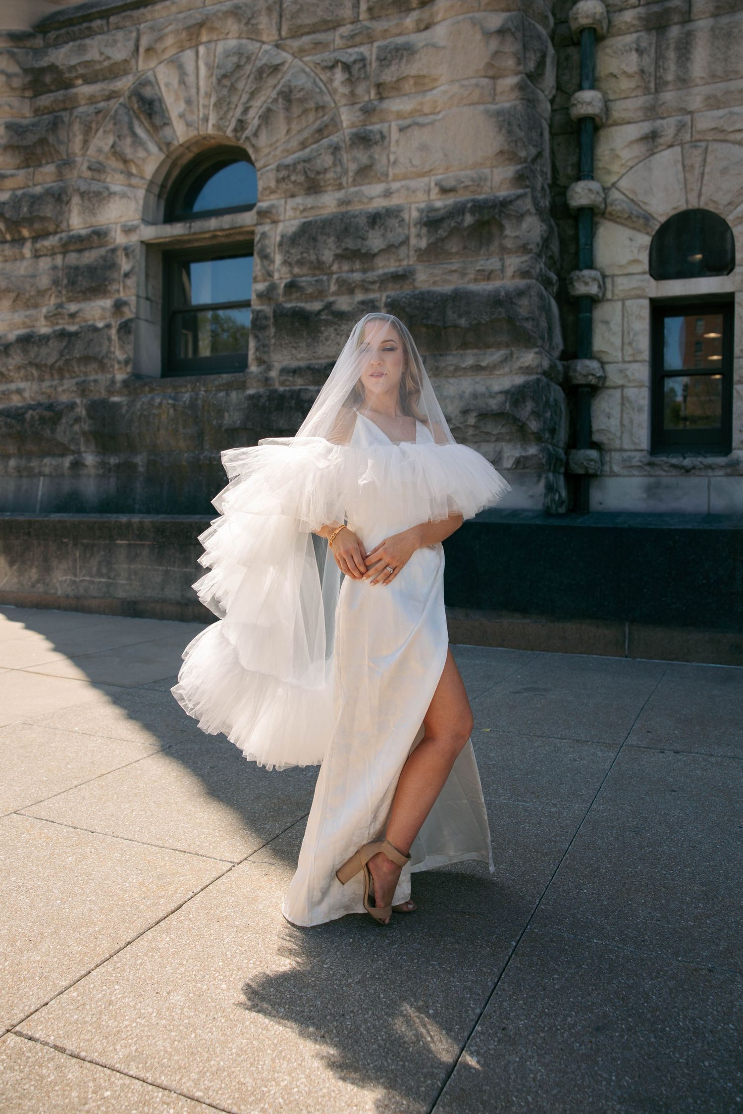 Bride in elegant white lace gown with layered tulle veil poses in golden sunlight outside historic stone building.