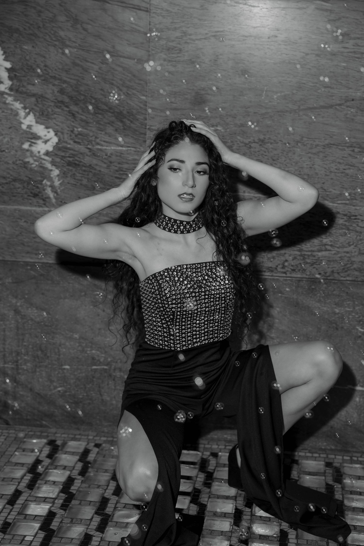 Black and white photo of a young woman in a studded corset and skirt, posing on a tiled floor with curly hair.