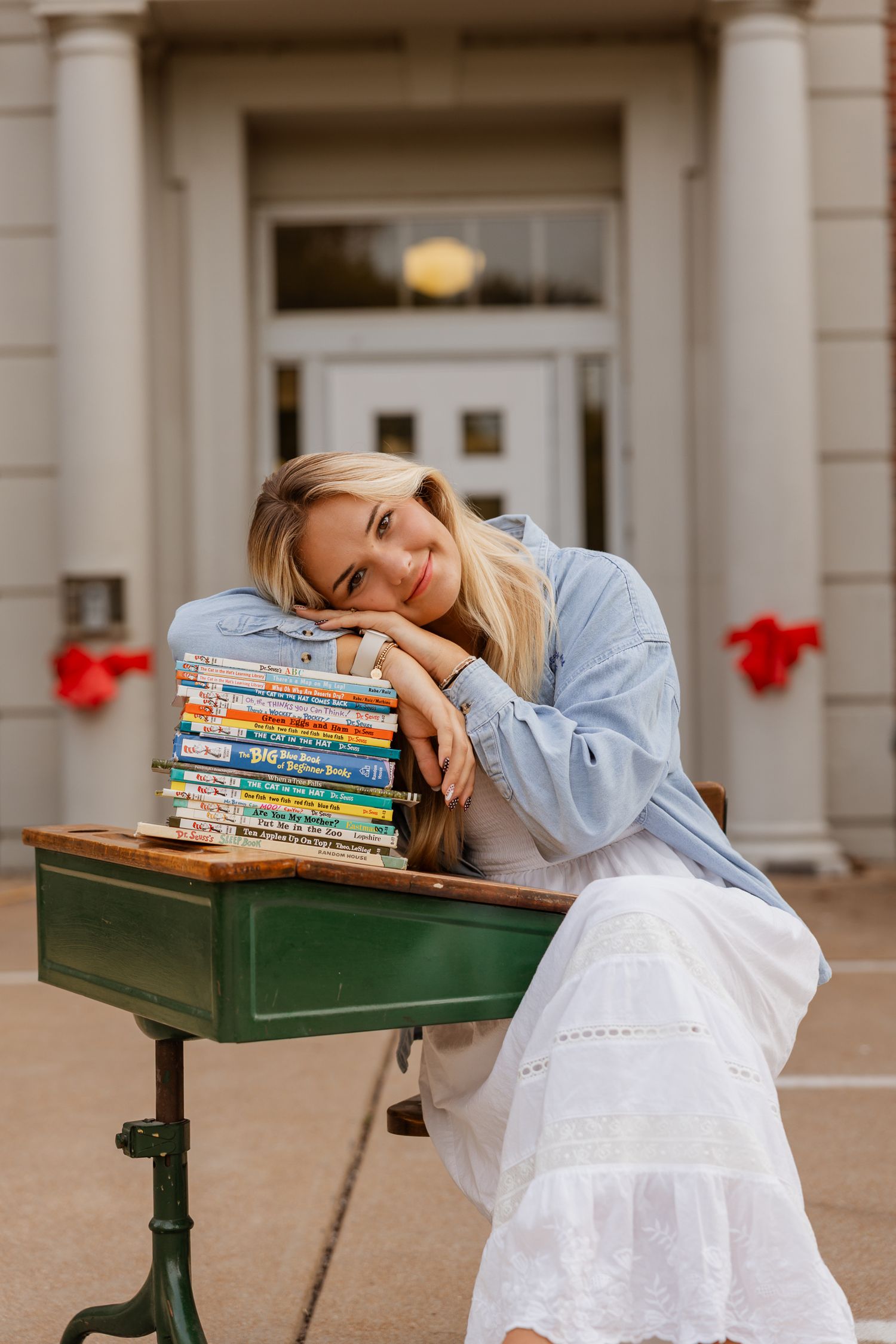 A collection of children's books stacked on a green desk outside a historic school building.
