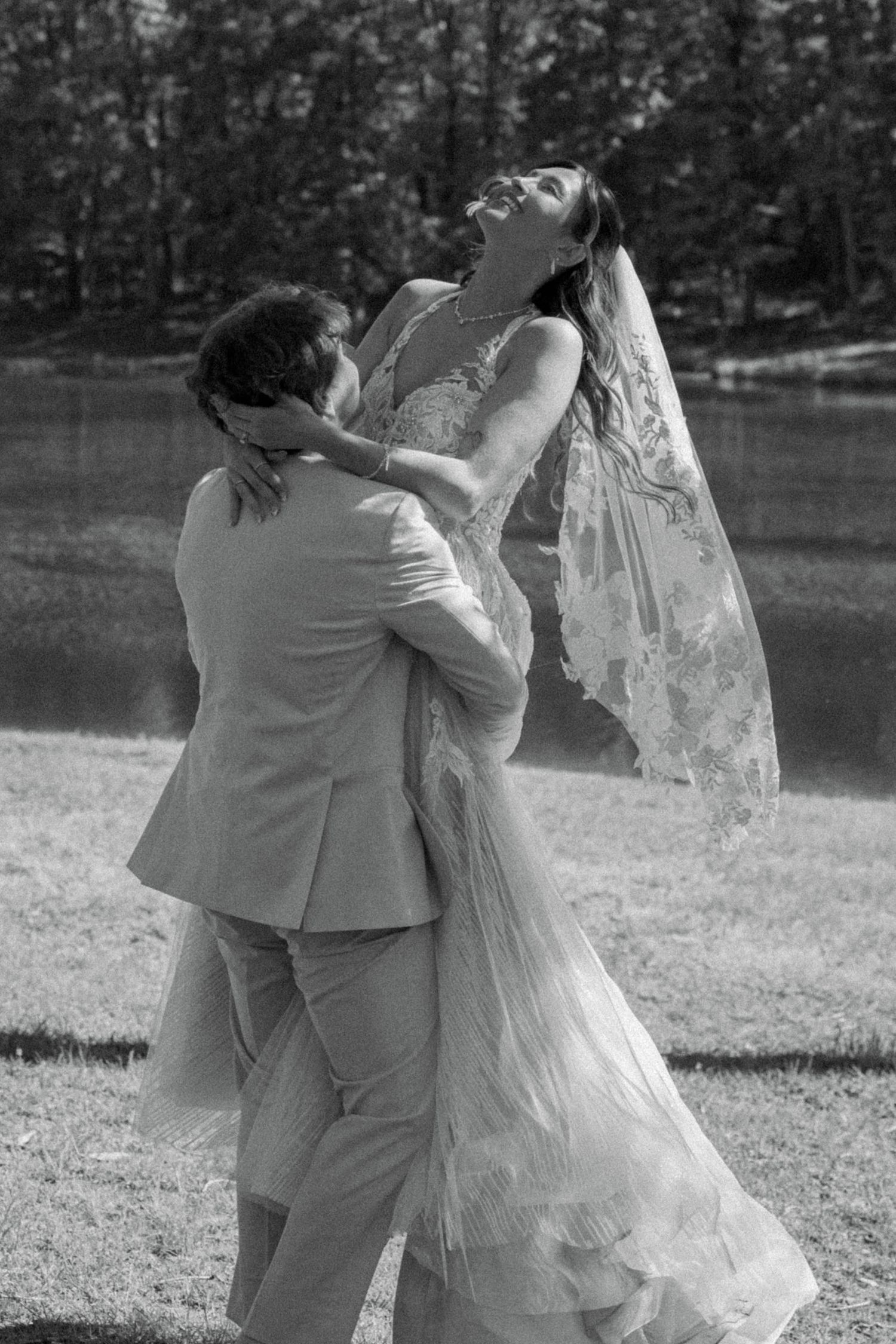 Black and white photo of a groom lifting his laughing bride in a lace wedding dress and veil outdoors.