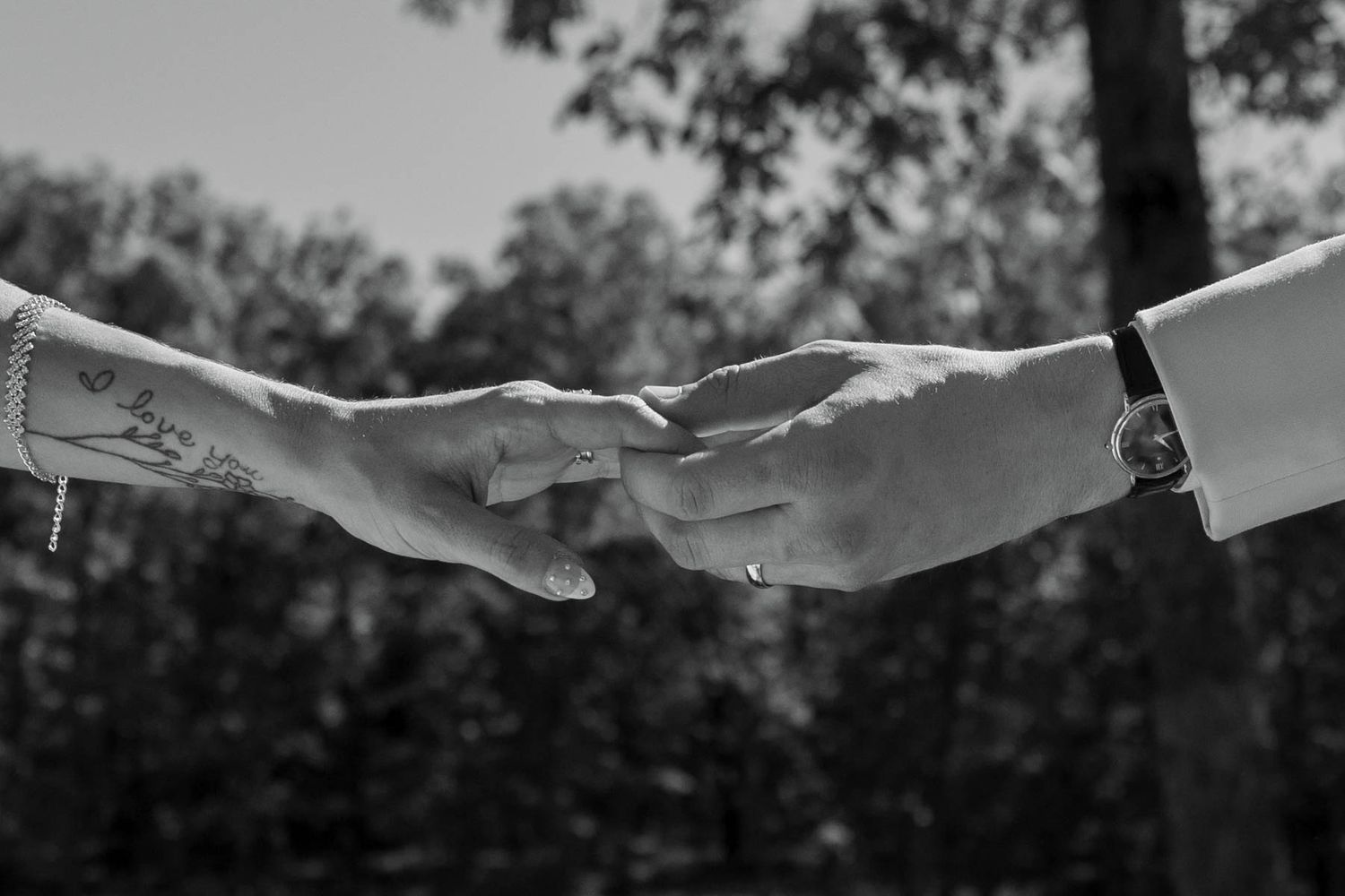 Black and white photo of two hands reaching toward each other, one with tattoos, exchanging rings outdoors.