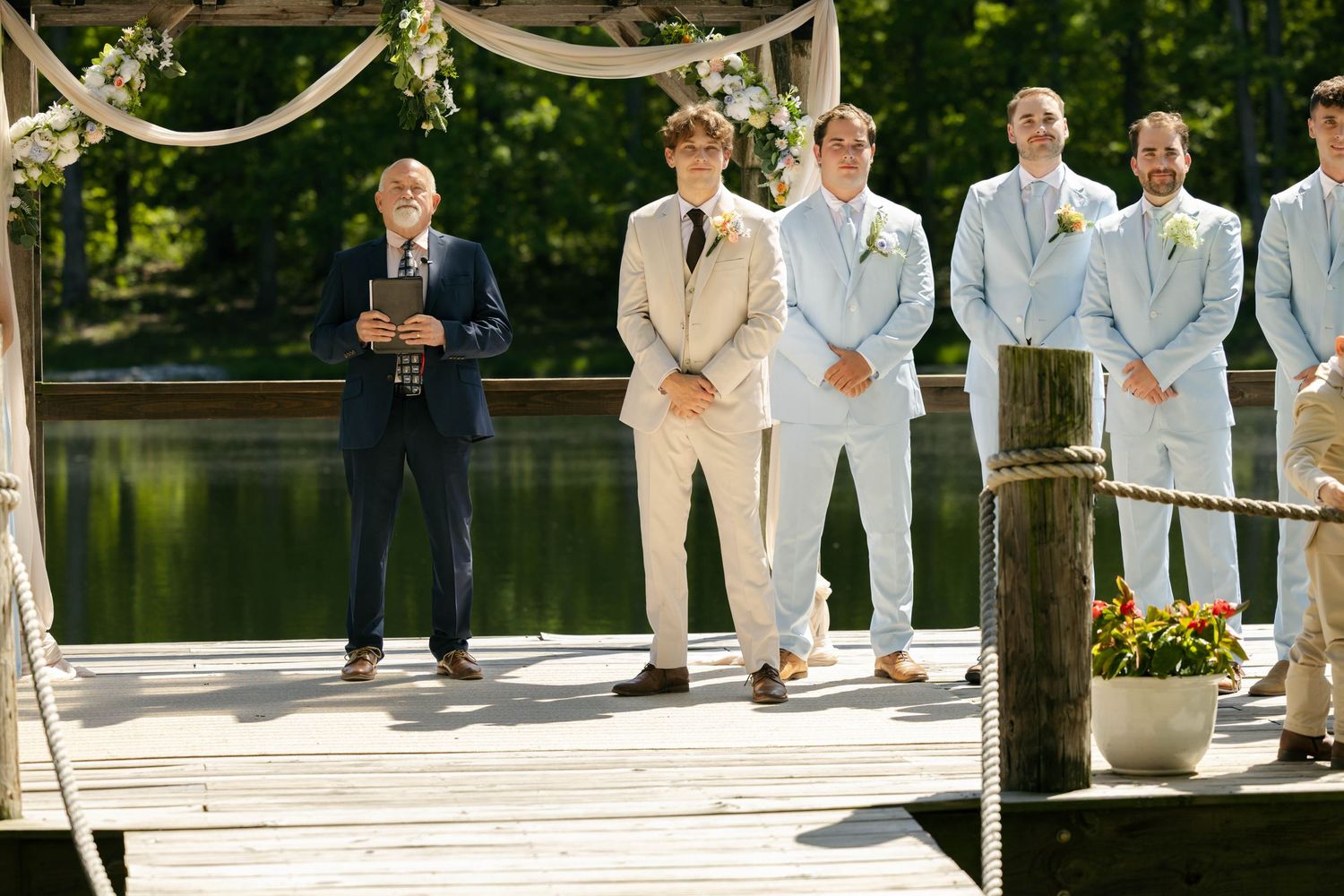 Groom and groomsmen in blue and tan suits stand on a lakeside dock during an outdoor wedding ceremony.