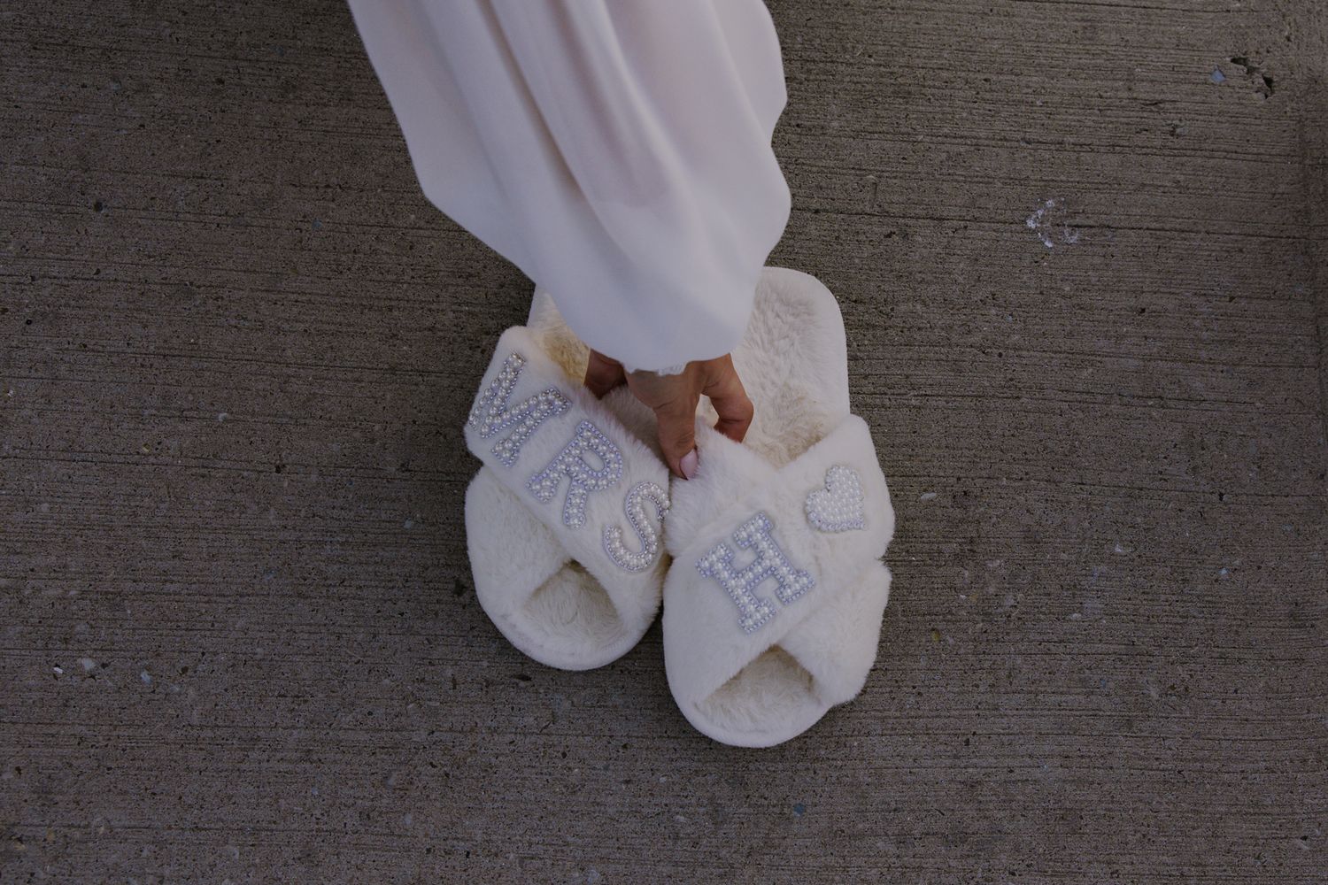 Person wearing white fluffy monogrammed slide slippers with cross-strap design, paired with white flowing pants.