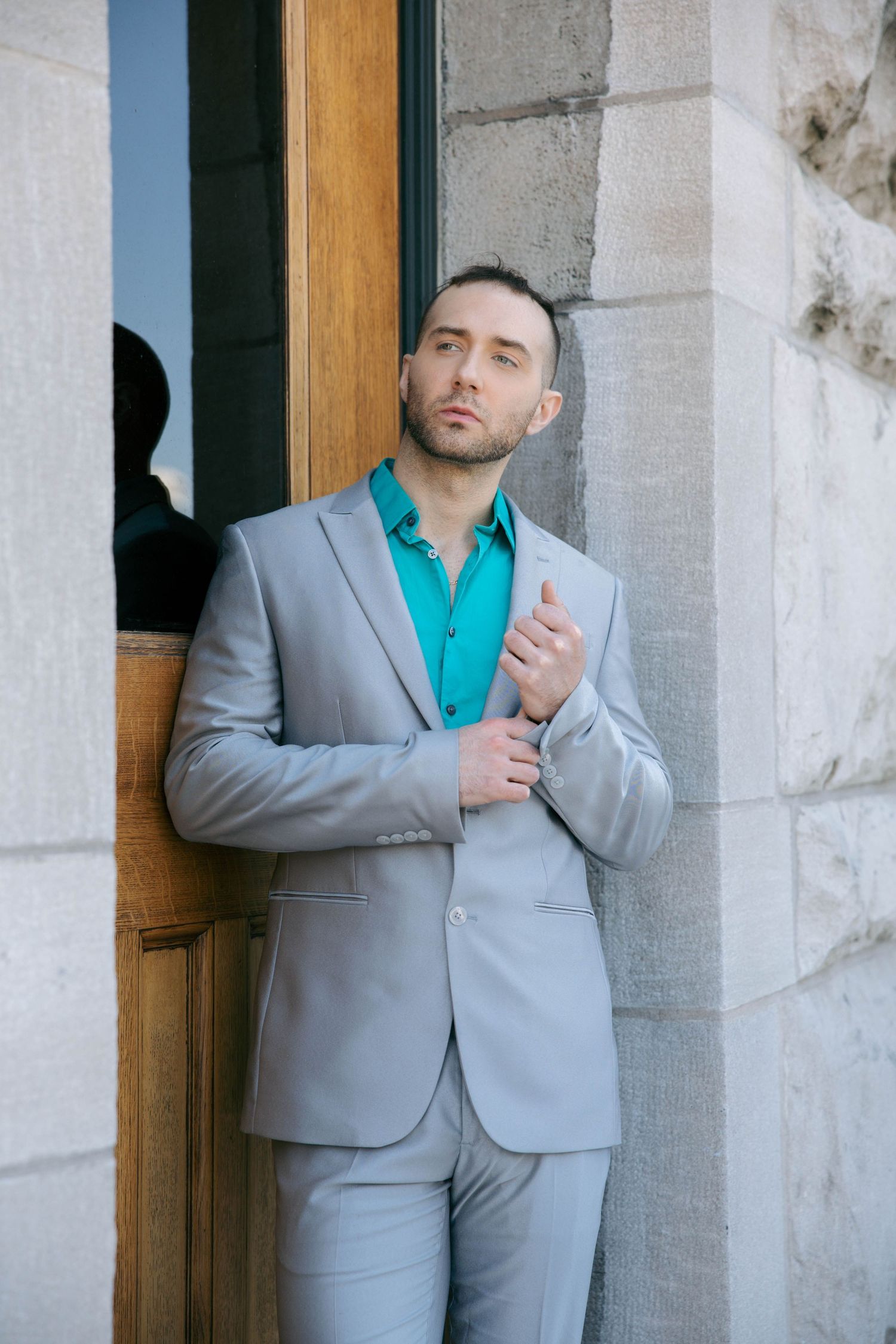 Stylish man in grey suit and teal shirt leans against stone doorway in a confident pose.
