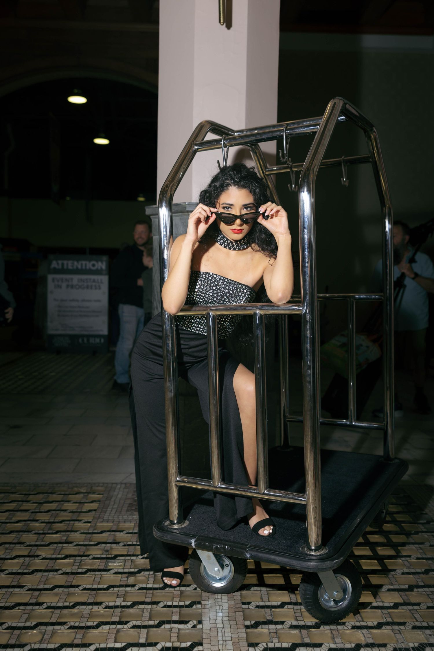 Stylish woman in black outfit standing inside hotel luggage cart adjusting sunglasses.