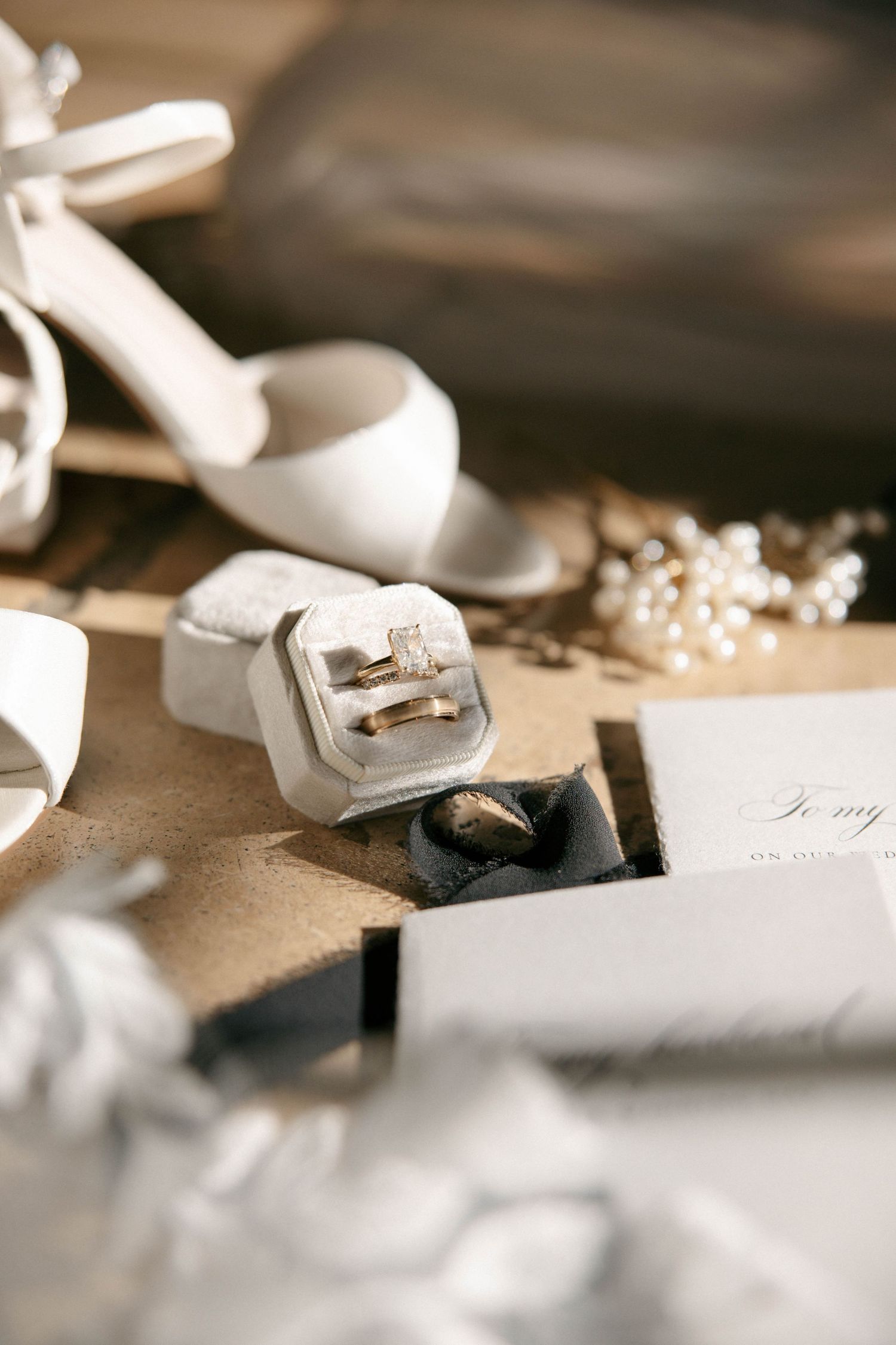 Wedding rings in a velvet box beside white heels, pearl necklace, and invitation on a wooden surface.