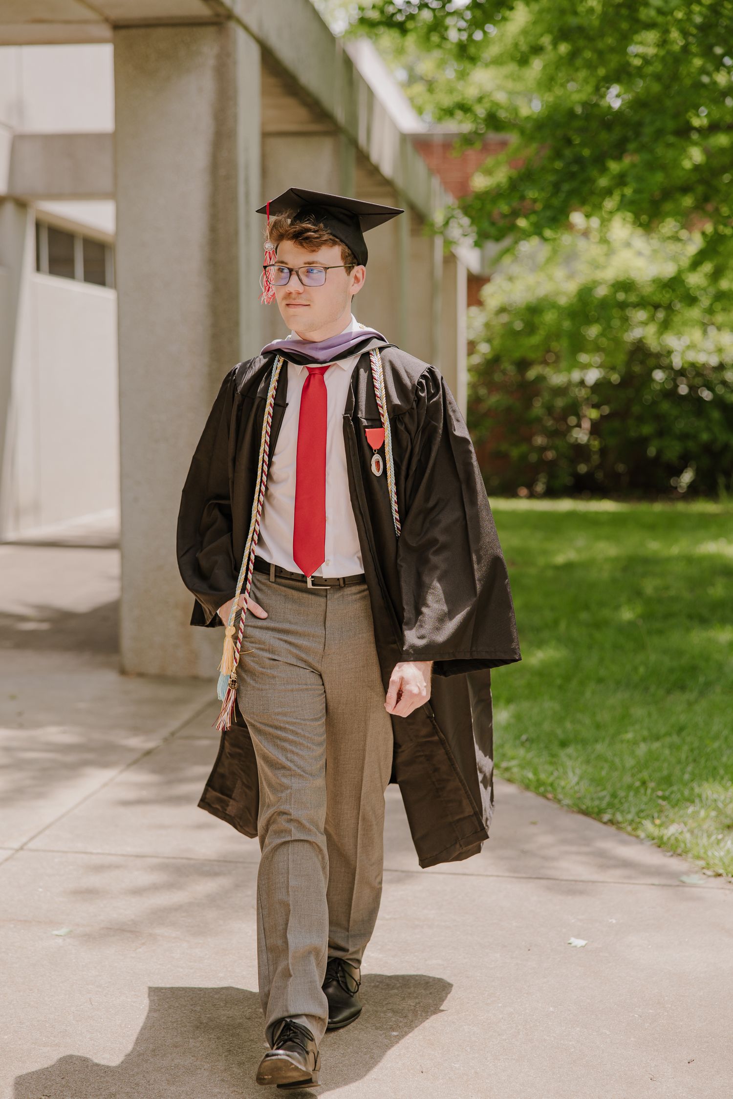 Graduate walks through campus colonnade wearing traditional black cap and gown with red tie.