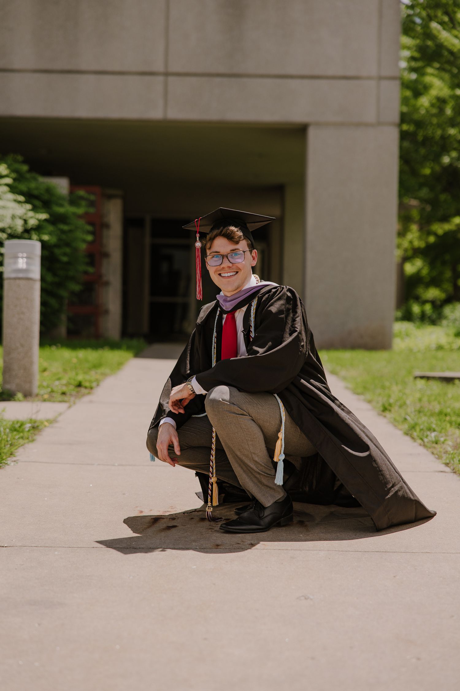 Graduate in cap and gown poses casually on concrete steps during outdoor graduation photoshoot.