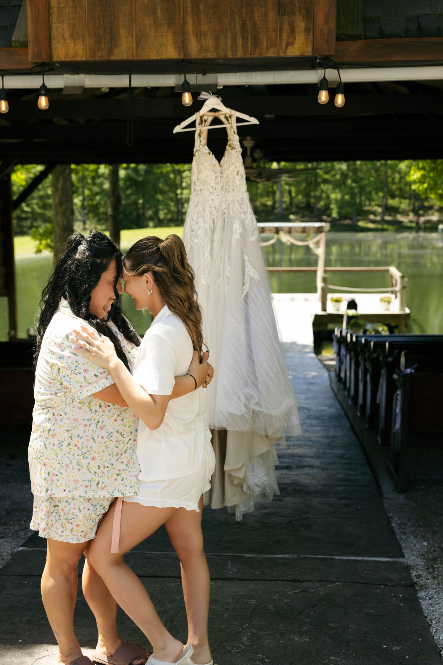 Two women embrace on a dock near a hanging white wedding dress at an outdoor waterfront venue.
