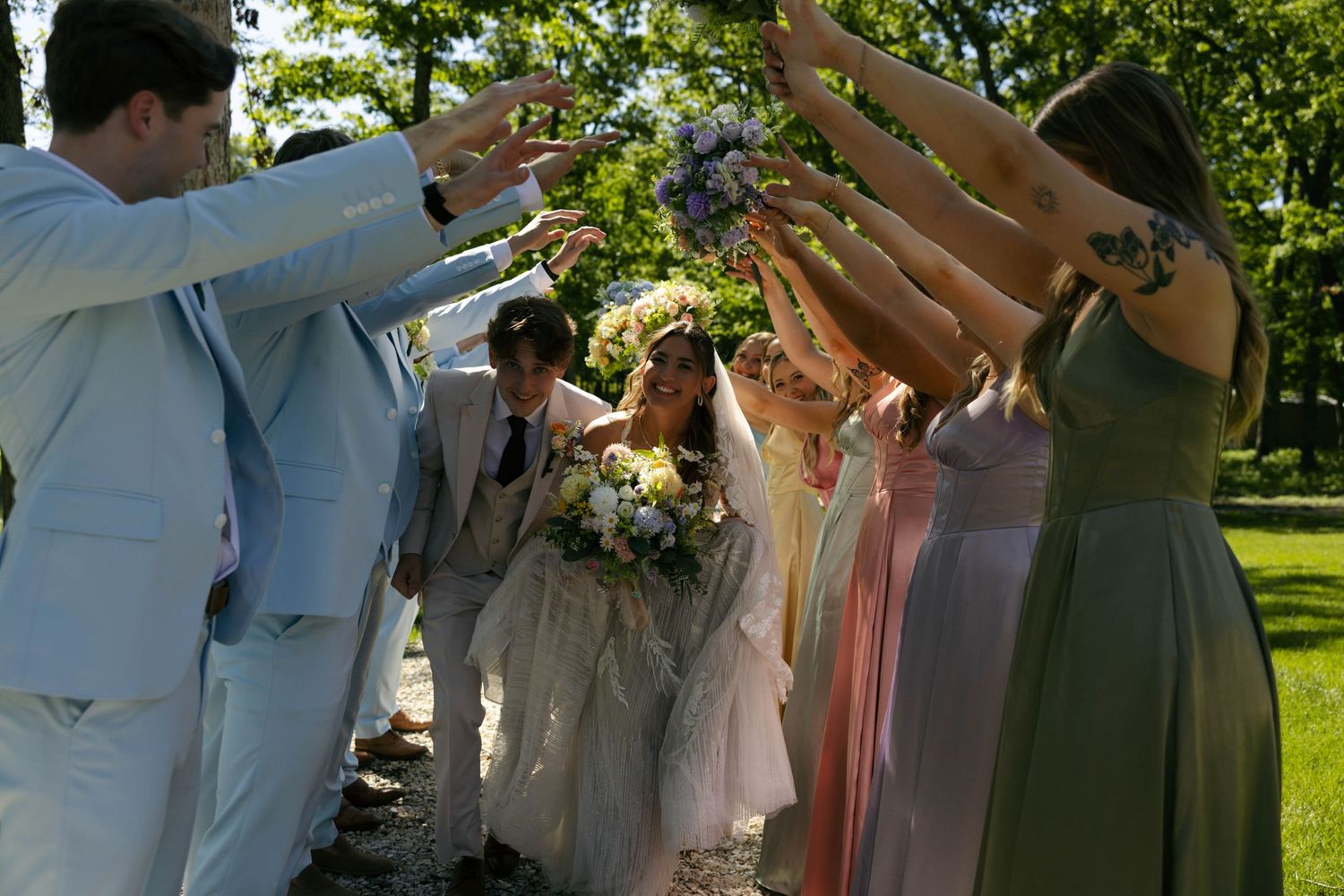 Wedding party celebrates newlyweds walking through an arch of raised hands outdoors in a sunlit garden.