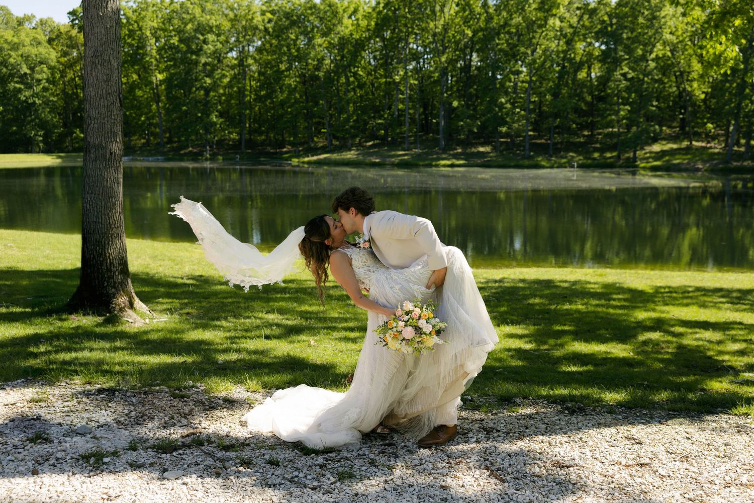 Bride and groom share a romantic dip kiss near a serene pond, with the veil flowing in the breeze on their wedding day.