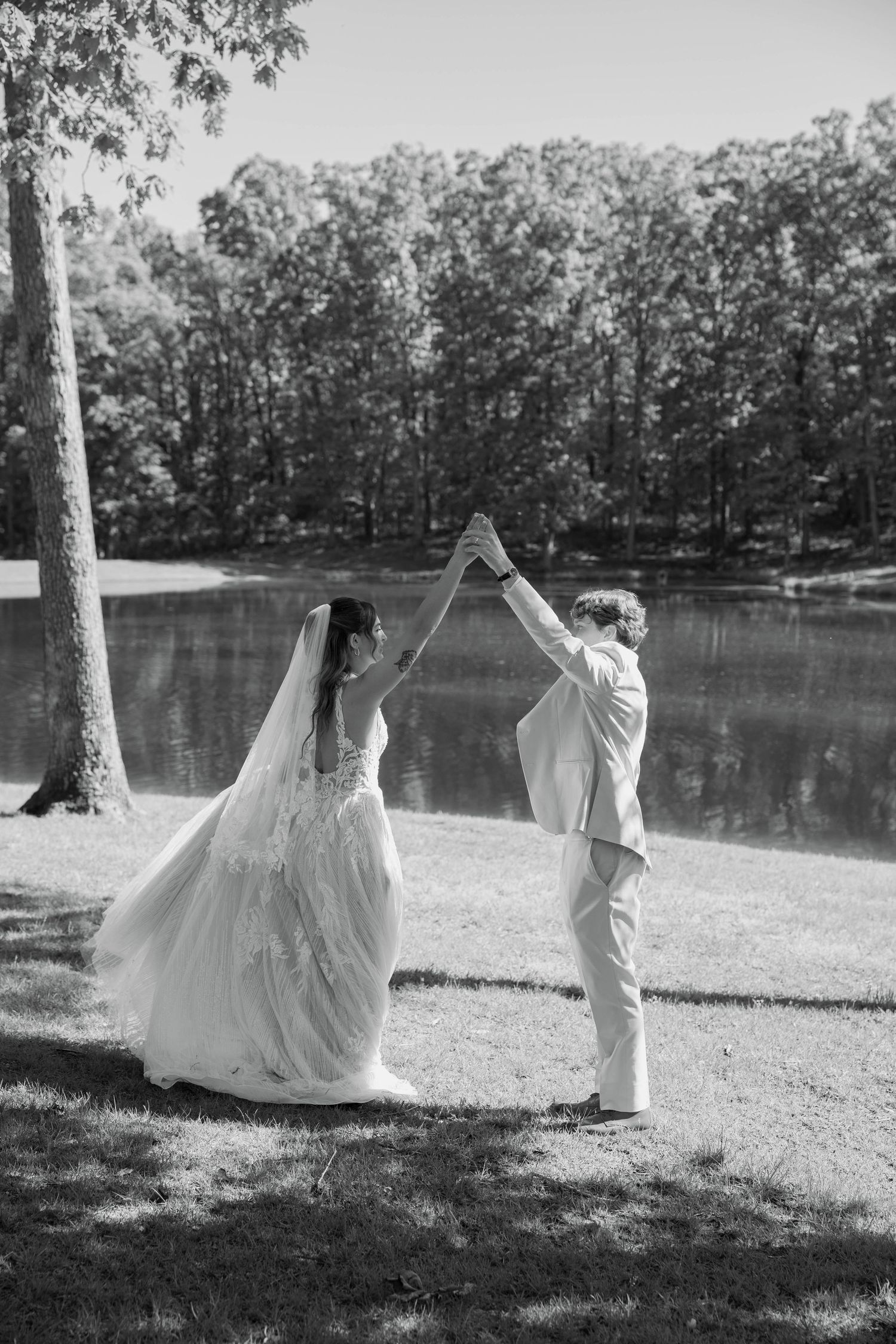 Black and white photo of bride and groom dancing by a lake, holding hands raised, surrounded by lush trees.
