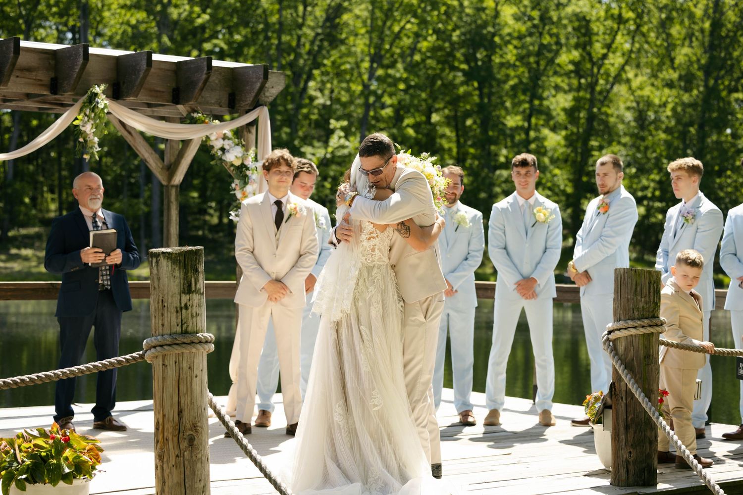 Groom lifting bride during outdoor waterfront wedding ceremony on wooden dock with groomsmen in light blue suits.