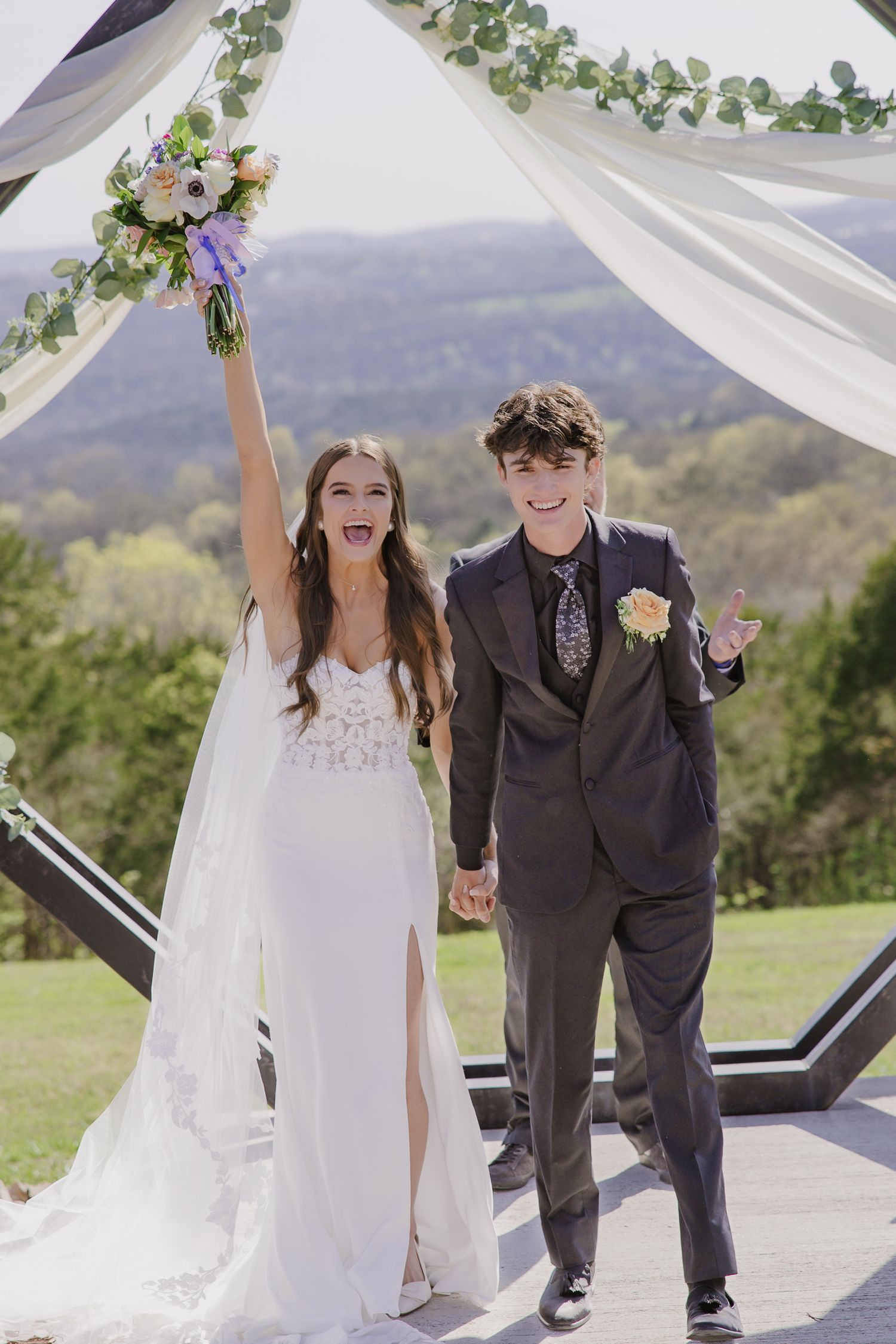 Newlyweds celebrate joyfully under a floral arch at an outdoor mountain venue.