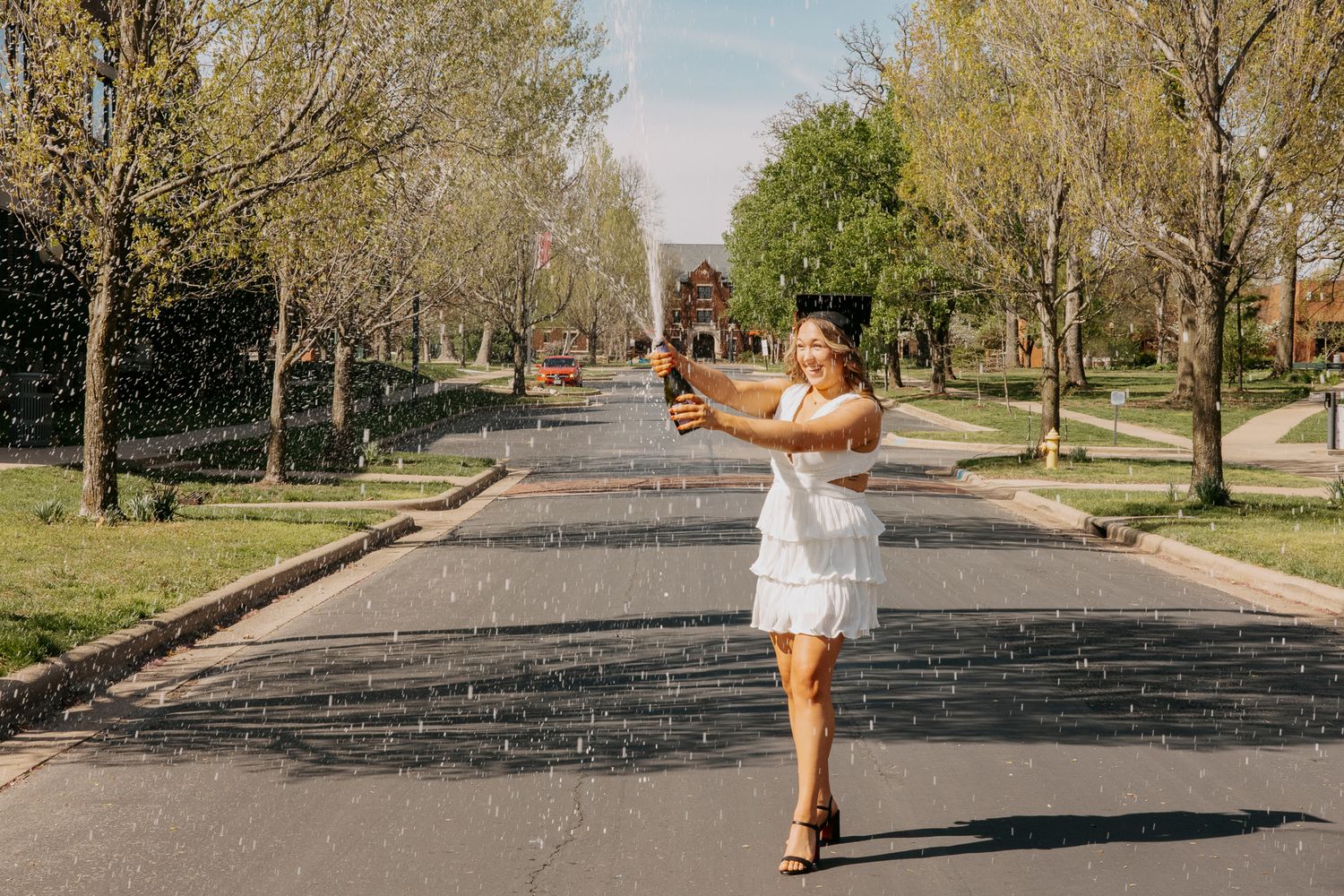 Graduate in white dress walking and posing on outdoor path surrounded by trees.