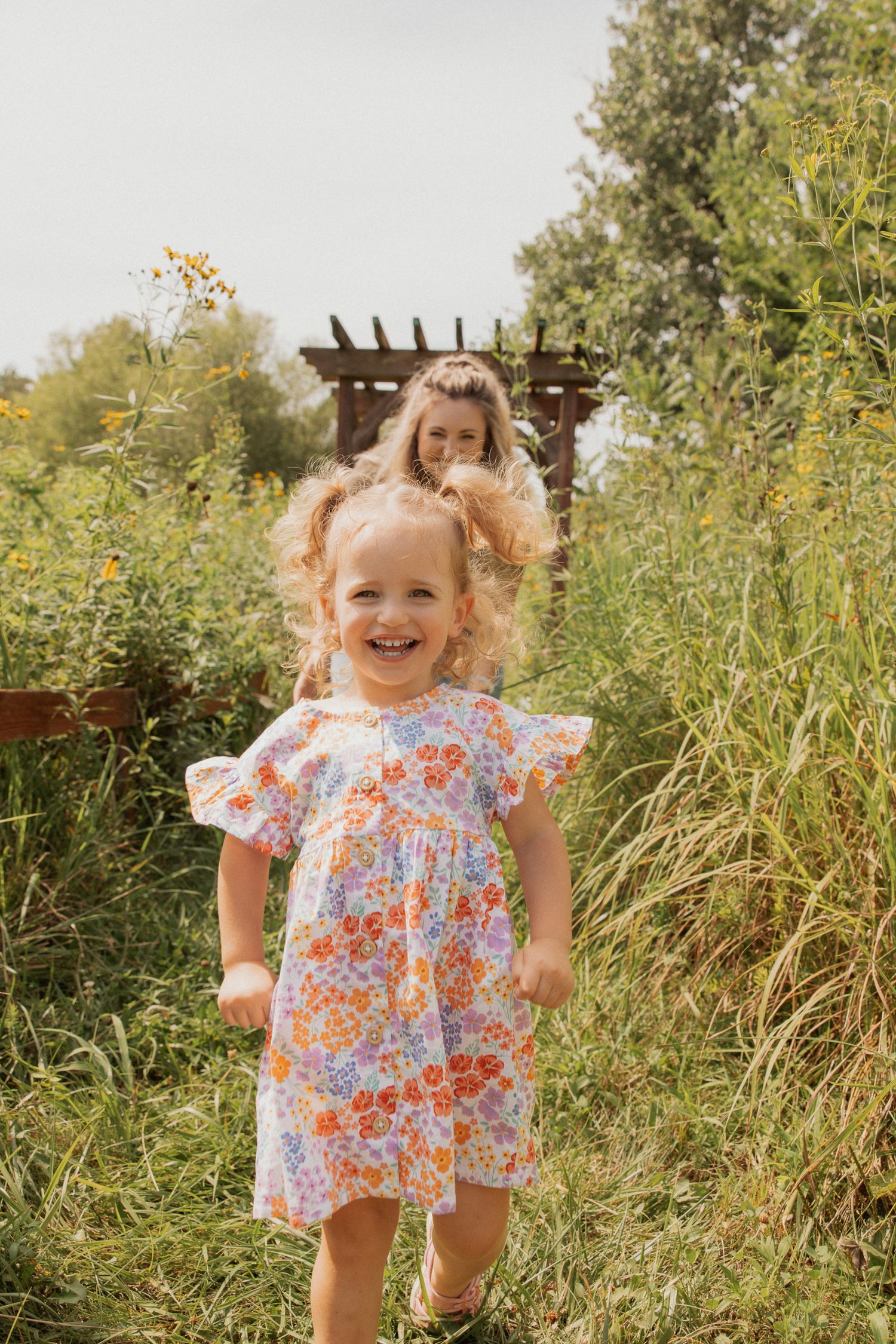 A floral dress twirls against a backdrop of summer greenery and a wooden pergola.