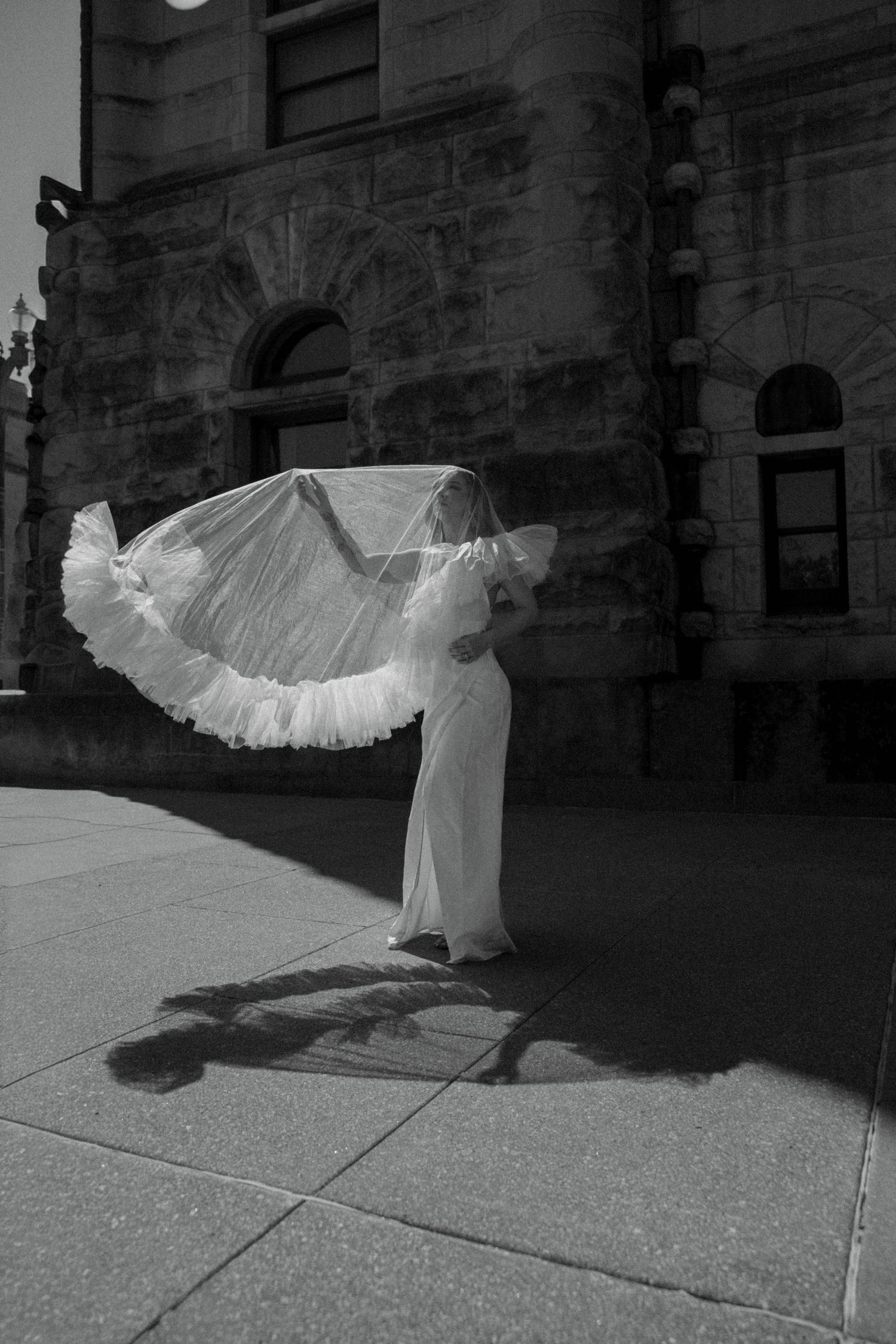 Black and white photo of bride in flowing white gown with billowing veil outside historic stone building.