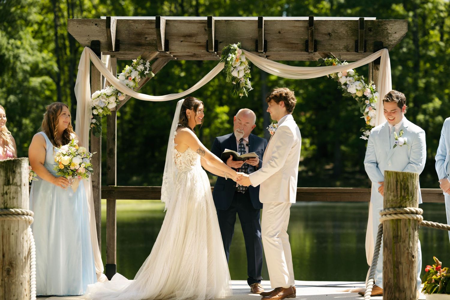 Outdoor wedding ceremony on a dock with bride and groom holding hands under a rustic floral arch by a serene lake.
