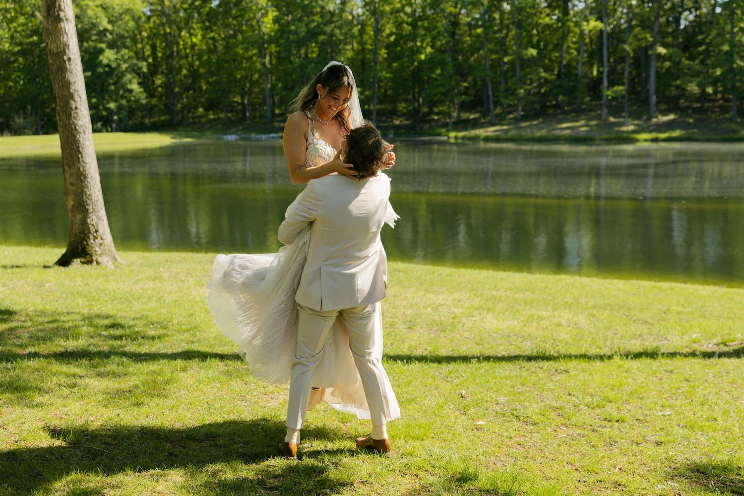 Groom lifting bride in white wedding dress by a sunlit pond surrounded by lush green trees.