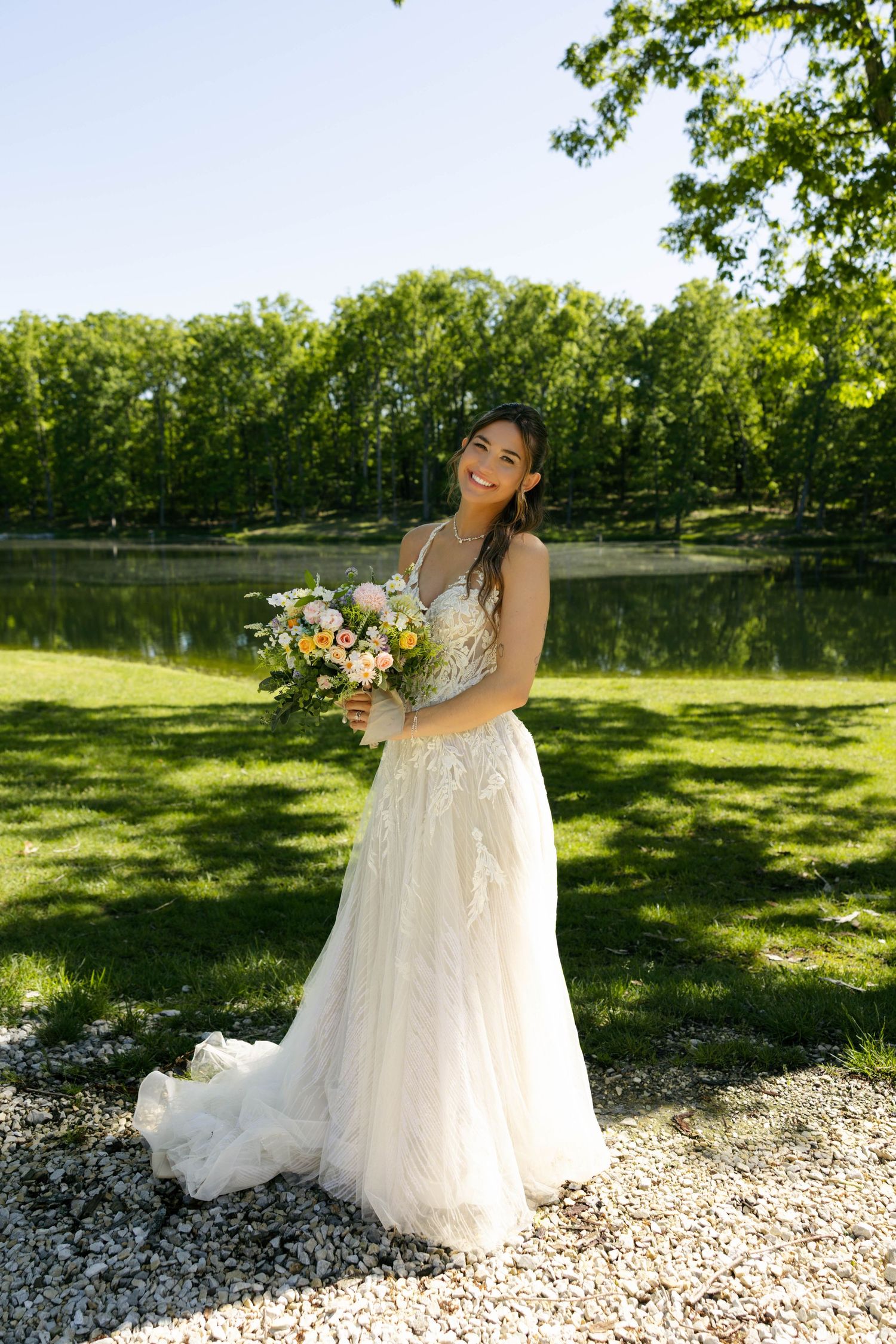 Smiling bride in white lace gown holding colorful bouquet outdoors near a pond surrounded by lush green trees.