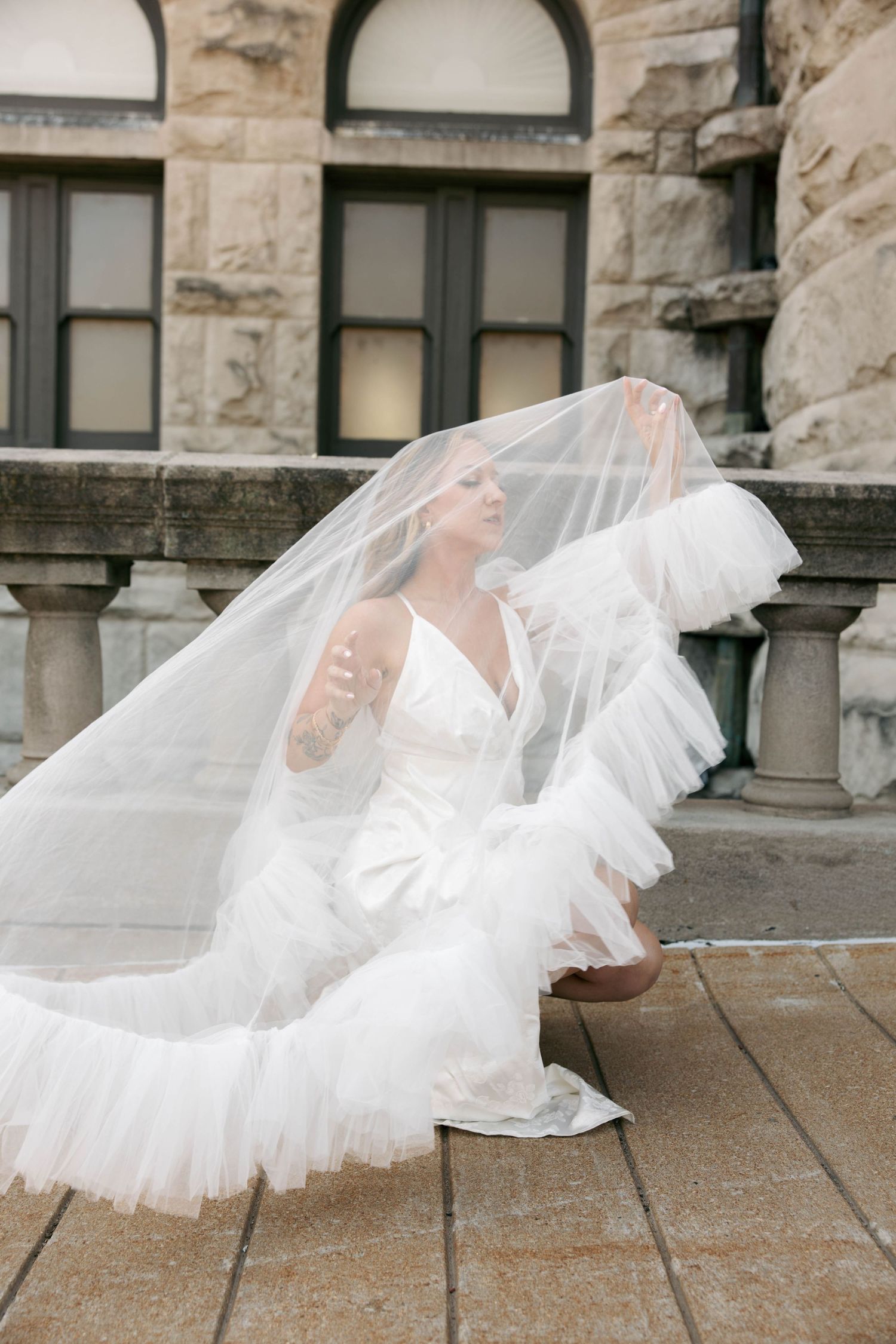 Bride in ruffled white gown crouches as her long veil billows around her near a stone building.