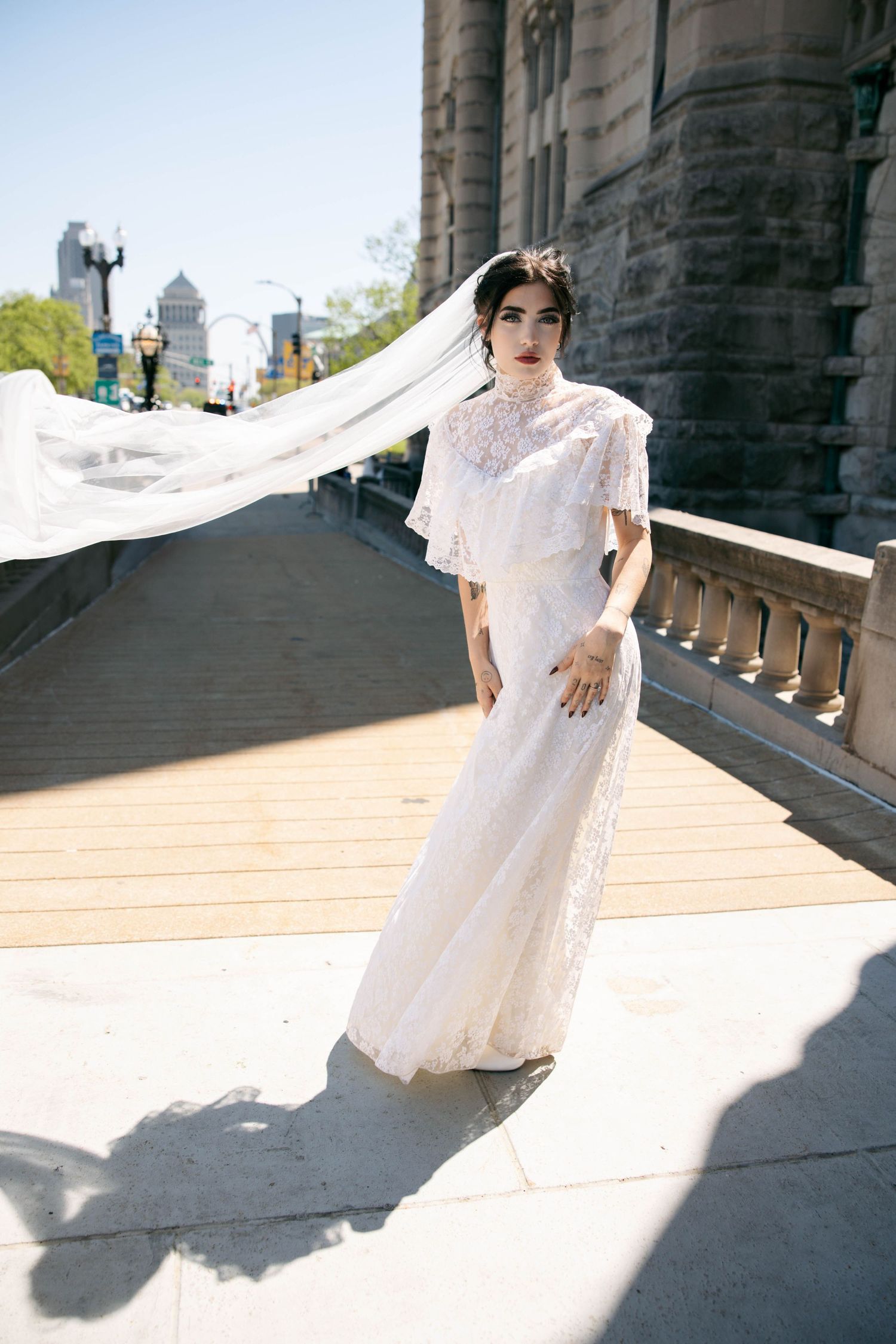 Bride in white lace high-neck gown with flowing veil outdoors on sunny urban steps.
